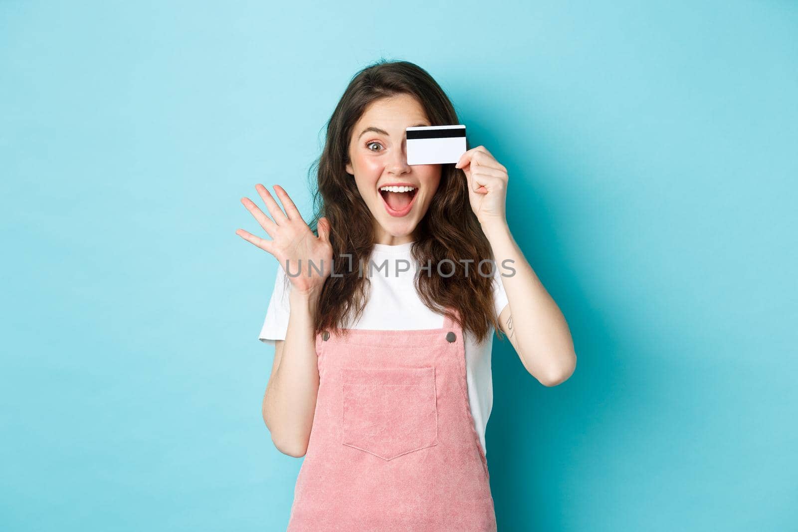 Excited and surprised young woman holding plastic credit card over eye, scream of joy while shopping, see something cool to buy, standing over blue background.