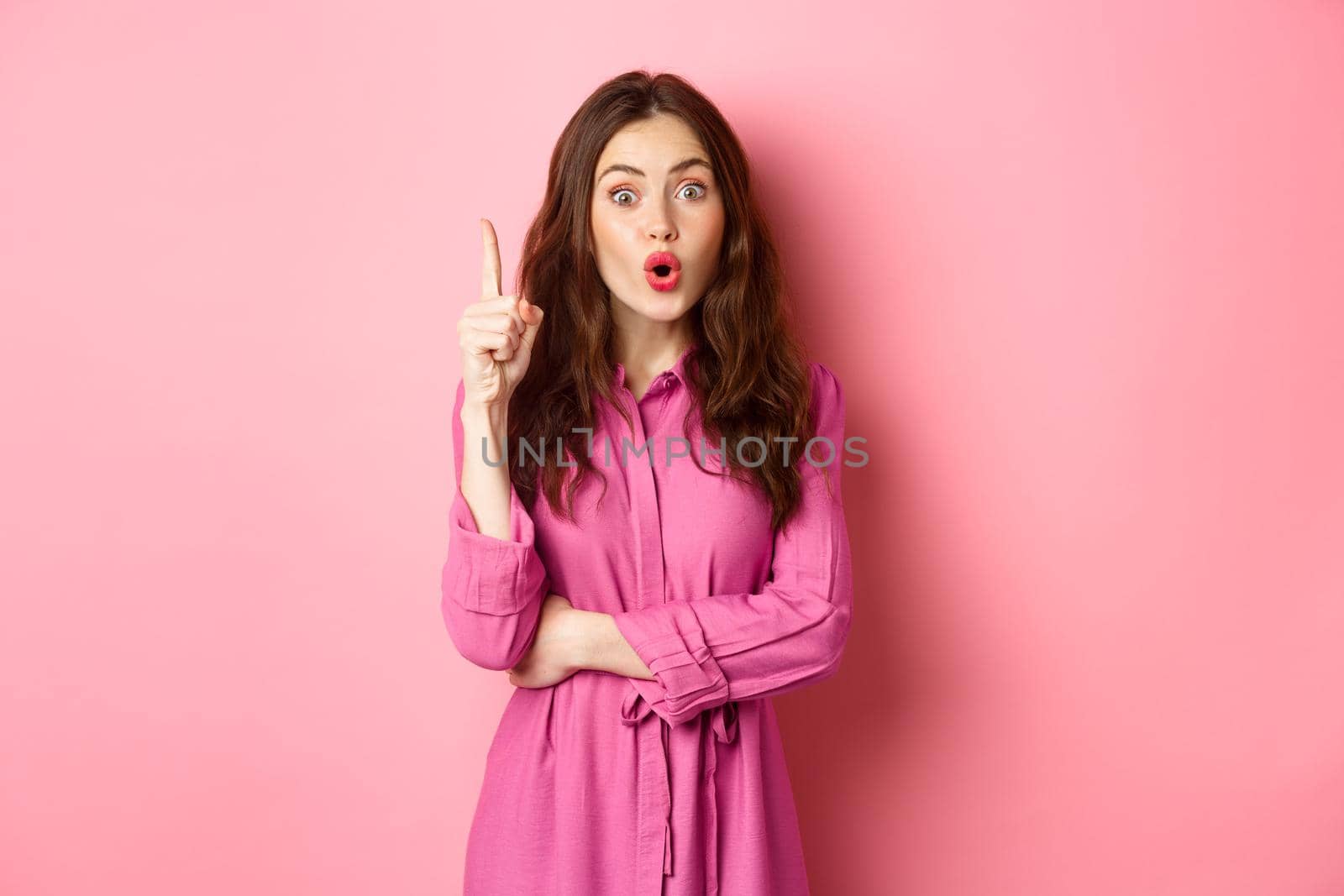 Young stylish feminine girl saying suggestion, have solution, got an idea, raising finger up in eureka sign, standing over pink background.
