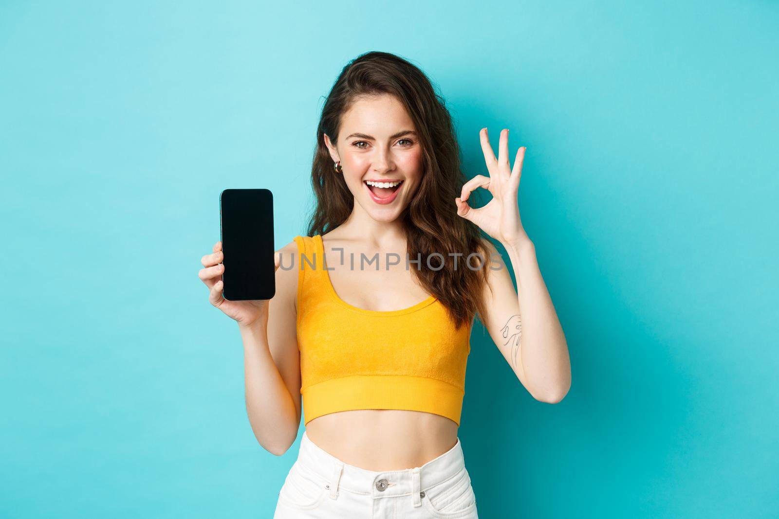 Look at this. Good-looking woman with sassy smile, winking and showing okay sign with empty smartphone screen, recommending app, standing over blue background by Benzoix