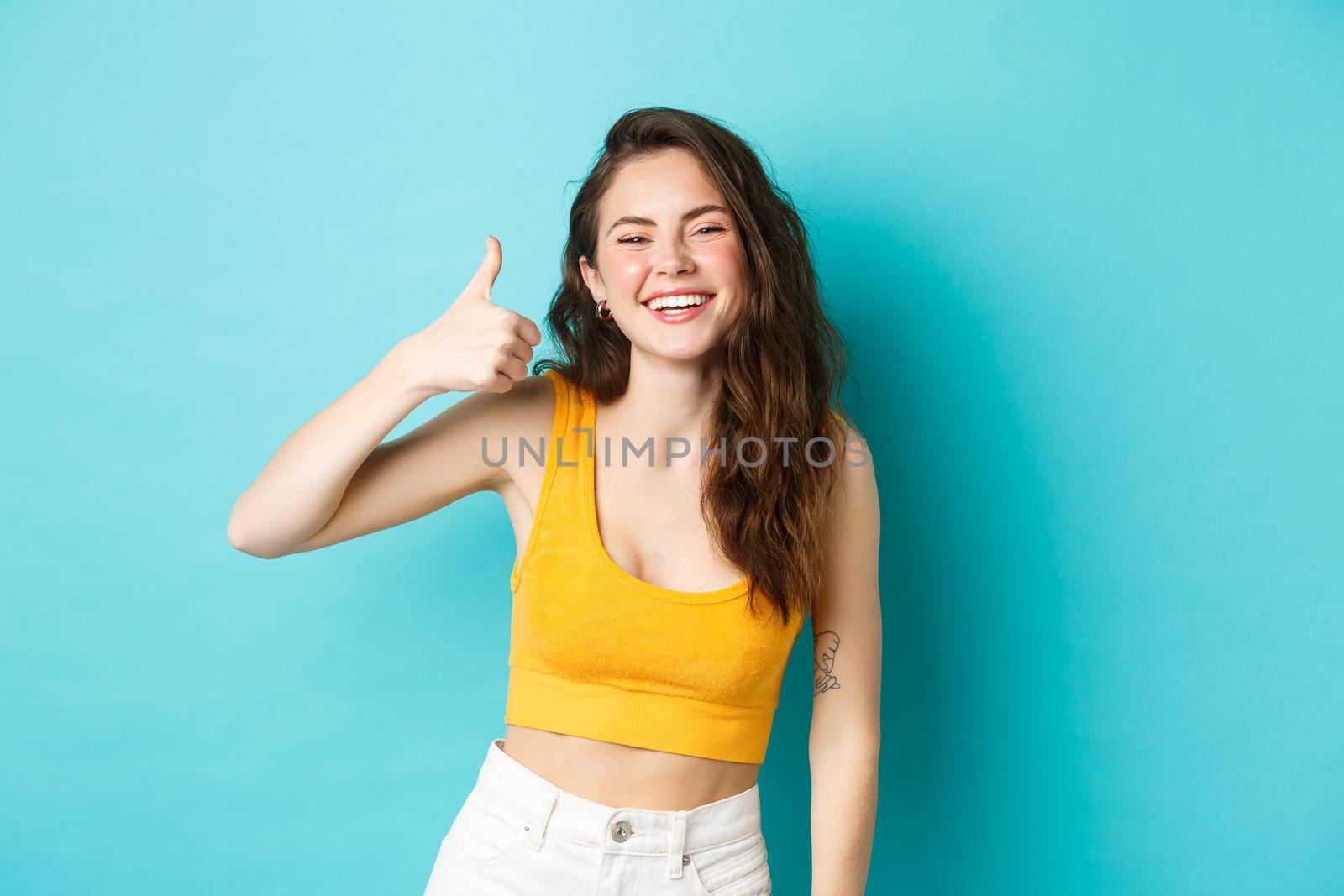 Summer holidays and emotions concept. Cheerful beauty girl with curly hairstyle and cropped top, showing yes thumb up gesture and laughing, standing over blue background.