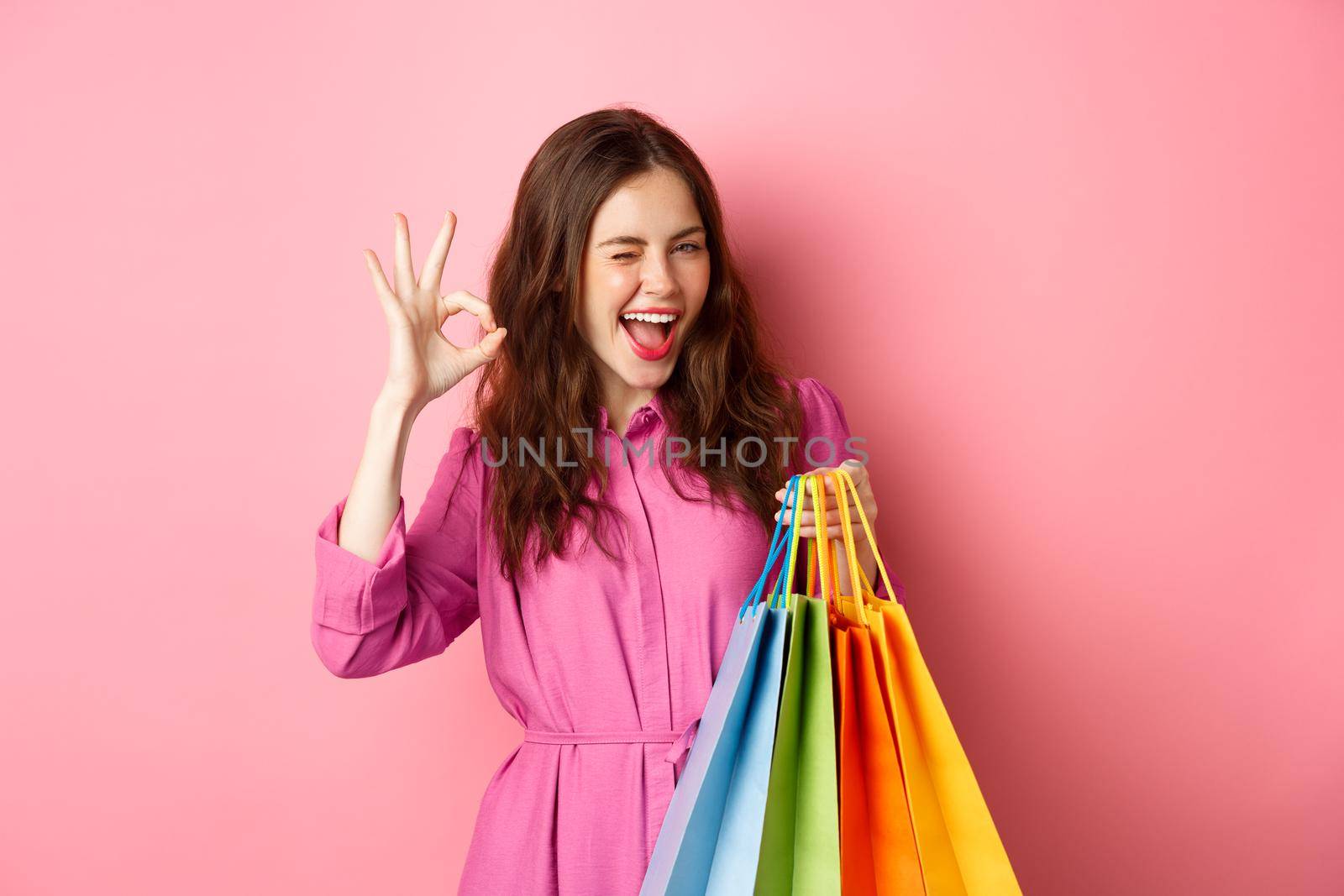 Young happy woman shopper showing okay sign, winking pleased with good discounts, buying staff on sale, holding shopping bags and smiling pleased, pink background.