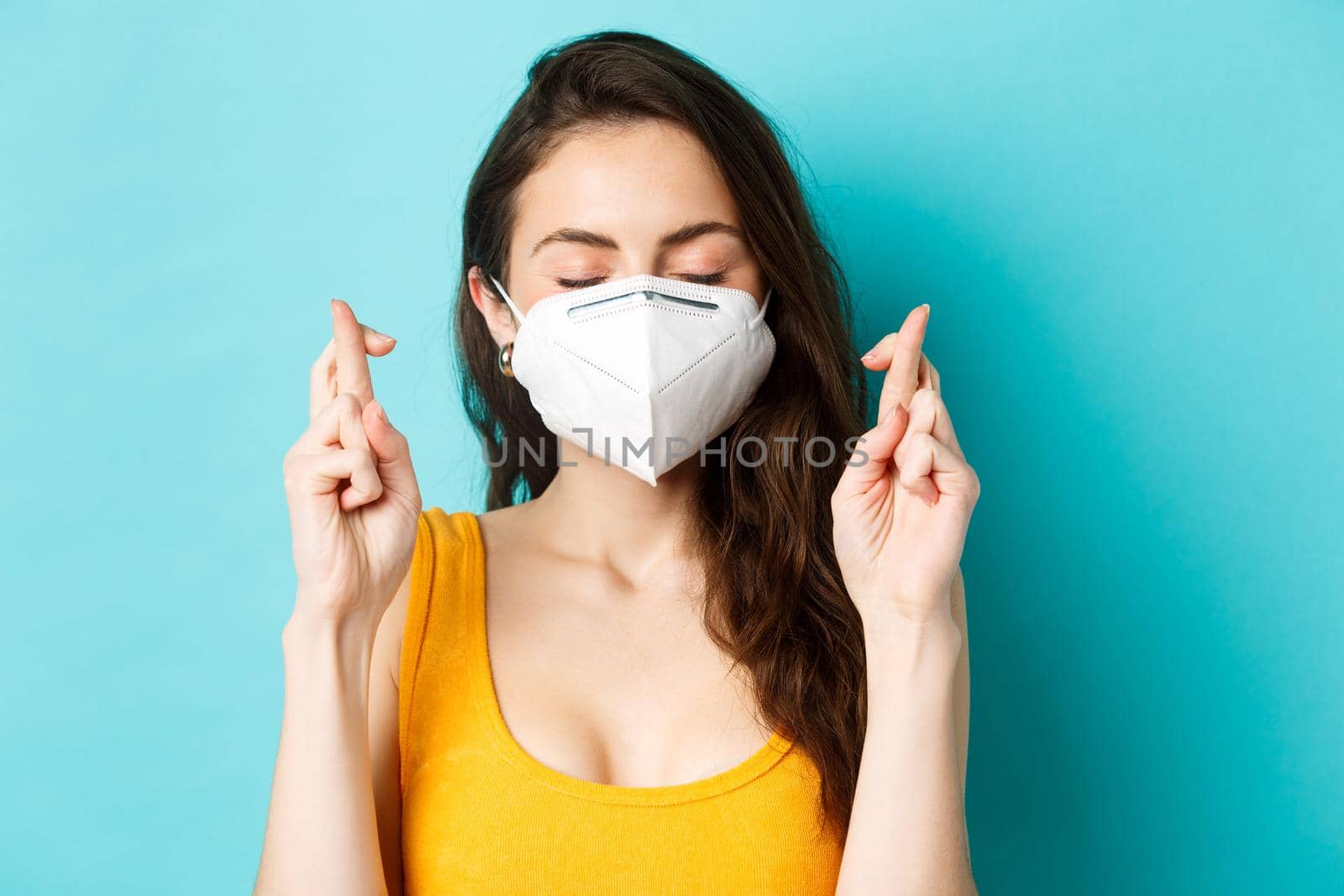 Health, covid-19 and lockdown concept. Close up of hopeful young woman in respirator mask, cross fingers and making wish, praying with eyes closed, standing against blue background.