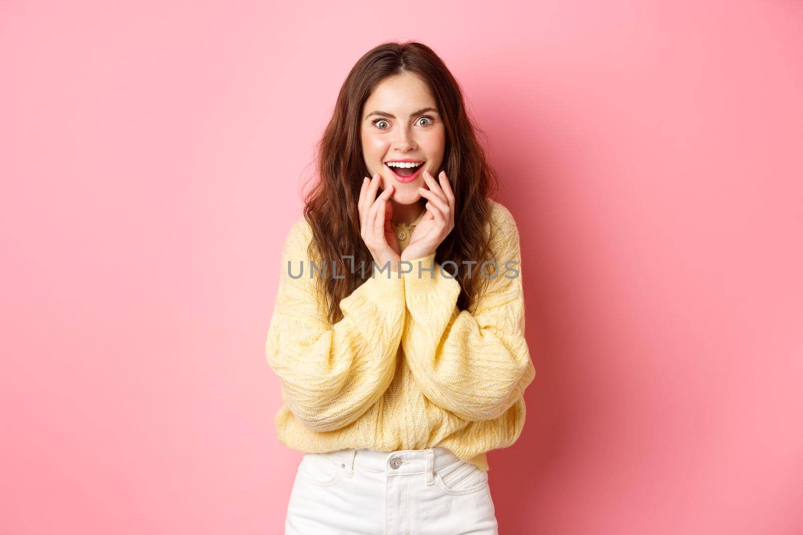 Omg really. Excited young woman hear gossip or super good news, staring amazed, smiling and holding hands on face, listen to interesting rumor, standing over pink background.