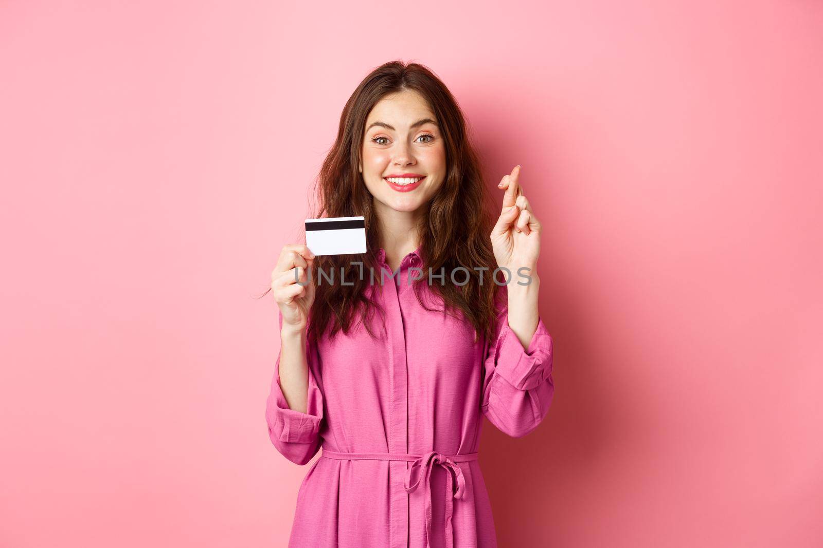 Image of hopeful young woman showing plastic credit card and cross fingers for good luck, making wish and smiling, standing against pink background.