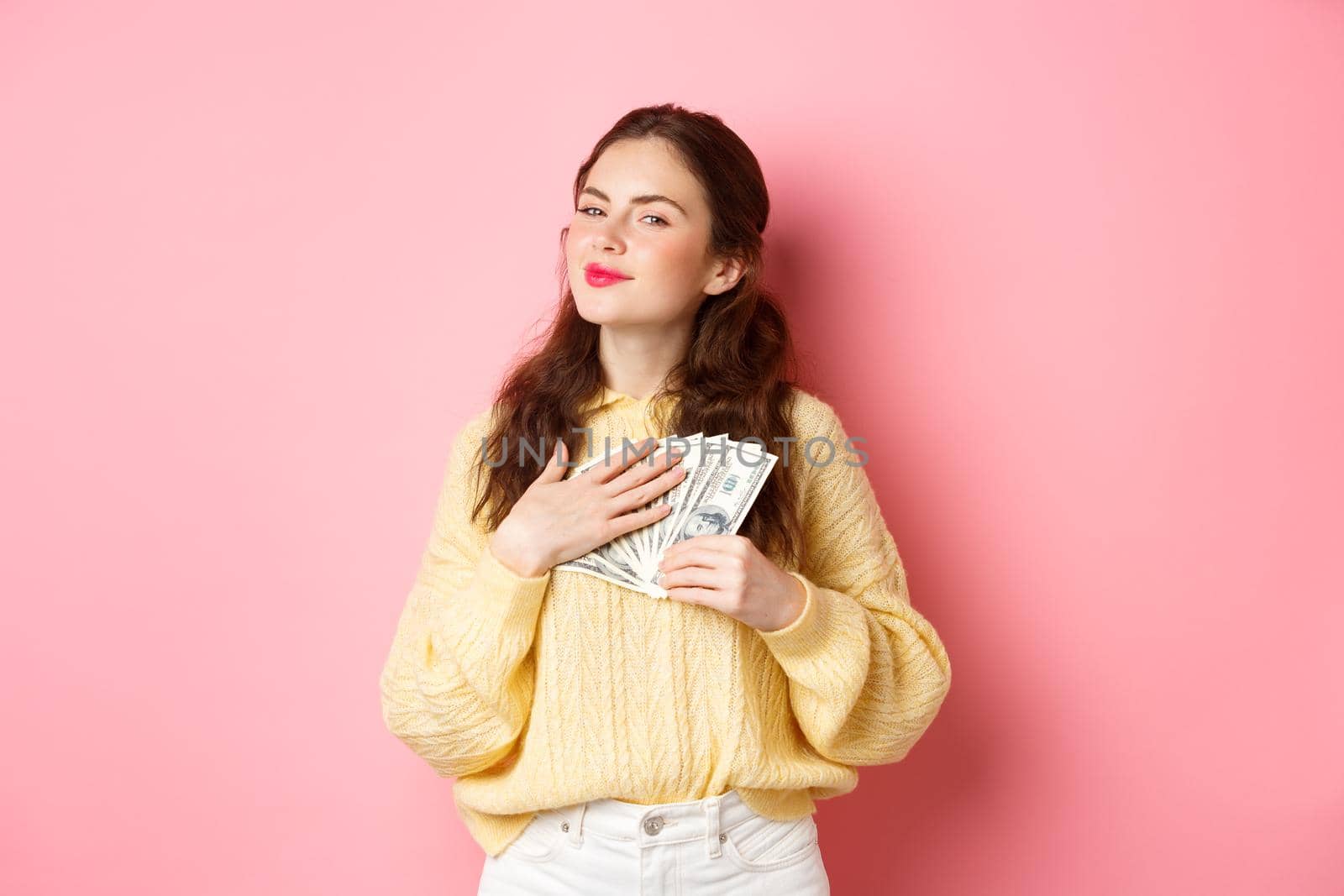 Smiling girl looks satisfied and grateful, hugging dollar bills, holding money and making smug face pleased, standing against pink background.