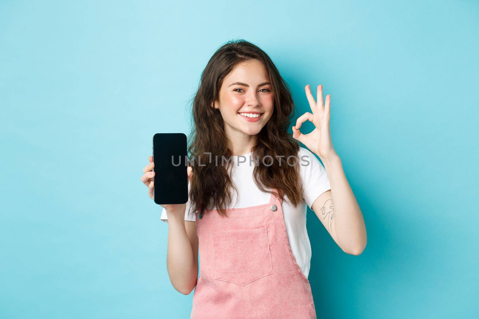 Very good. Smiling young woman showing product online on empty smartphone screen, recommending your brand, make ok sign, standing satisfied against blue background.