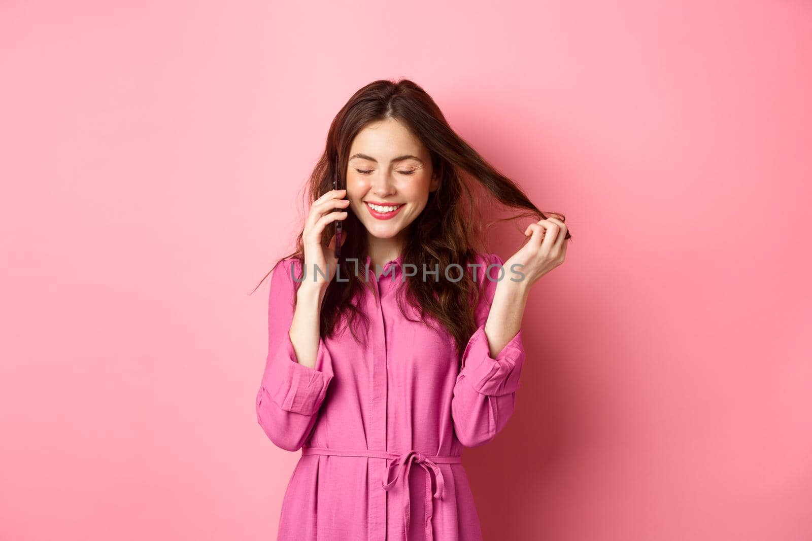 Beautiful young woman calling someone, laughing and smiling during phone call, talking to friend and playing with hair strands, standing against pink background.