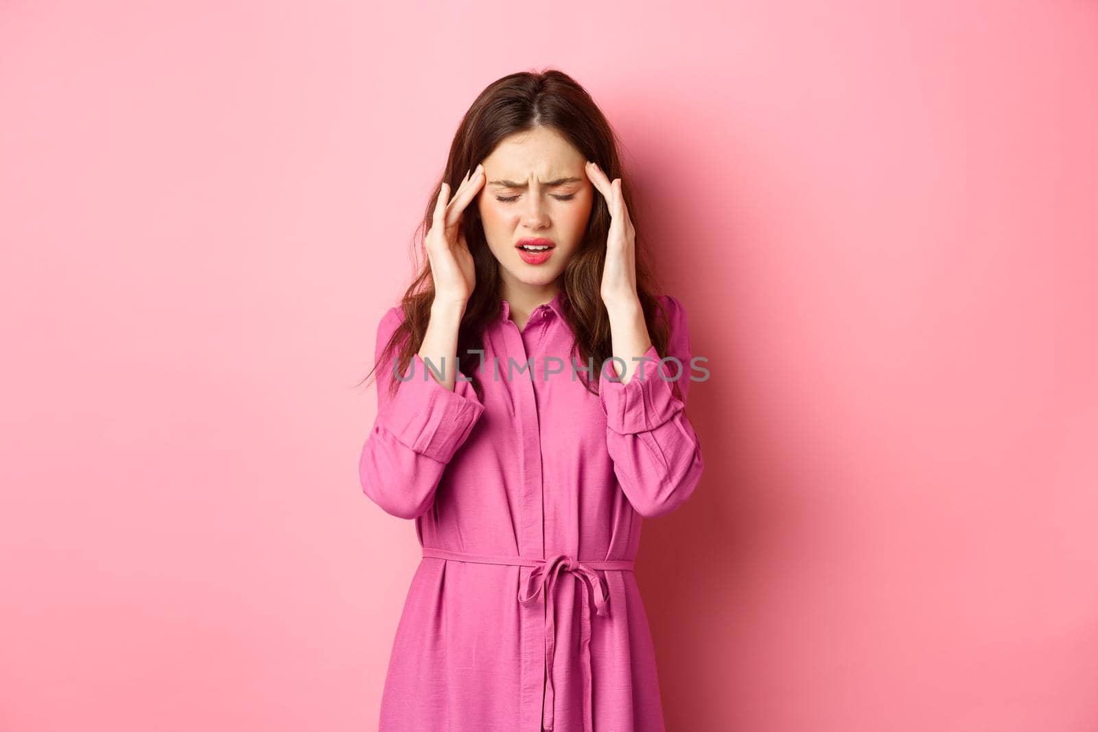 Woman feeling pain in head, touching temples and frowning, suffering migraine, being dizzy, standing with headache against pink background by Benzoix