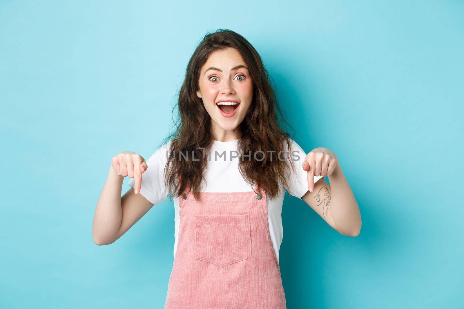 Wow so awesome. Excited cute girl telling big news, smiling amazed and pointing fingers down, showing cool advertisement, giving advice, recommending check out promo.