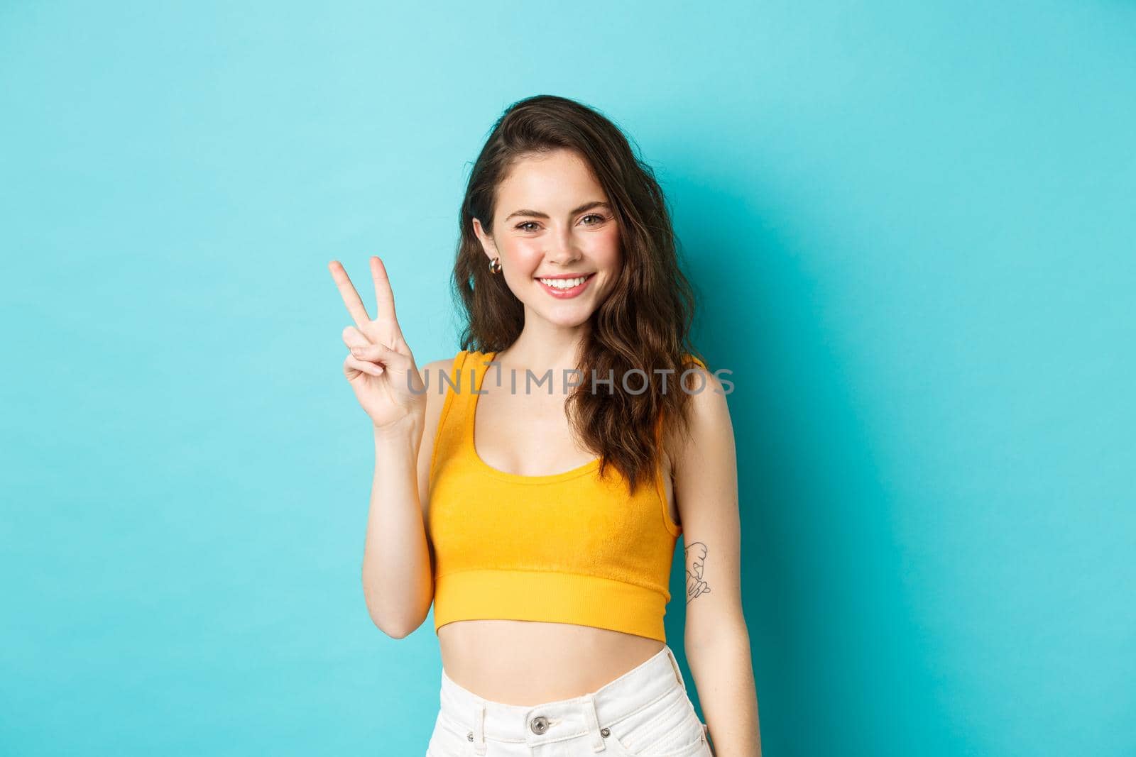 Summer and lifestyle concept. Beautiful girl tourist in crop top, posing with v-sign and smiling happy at camera, standing against blue background.