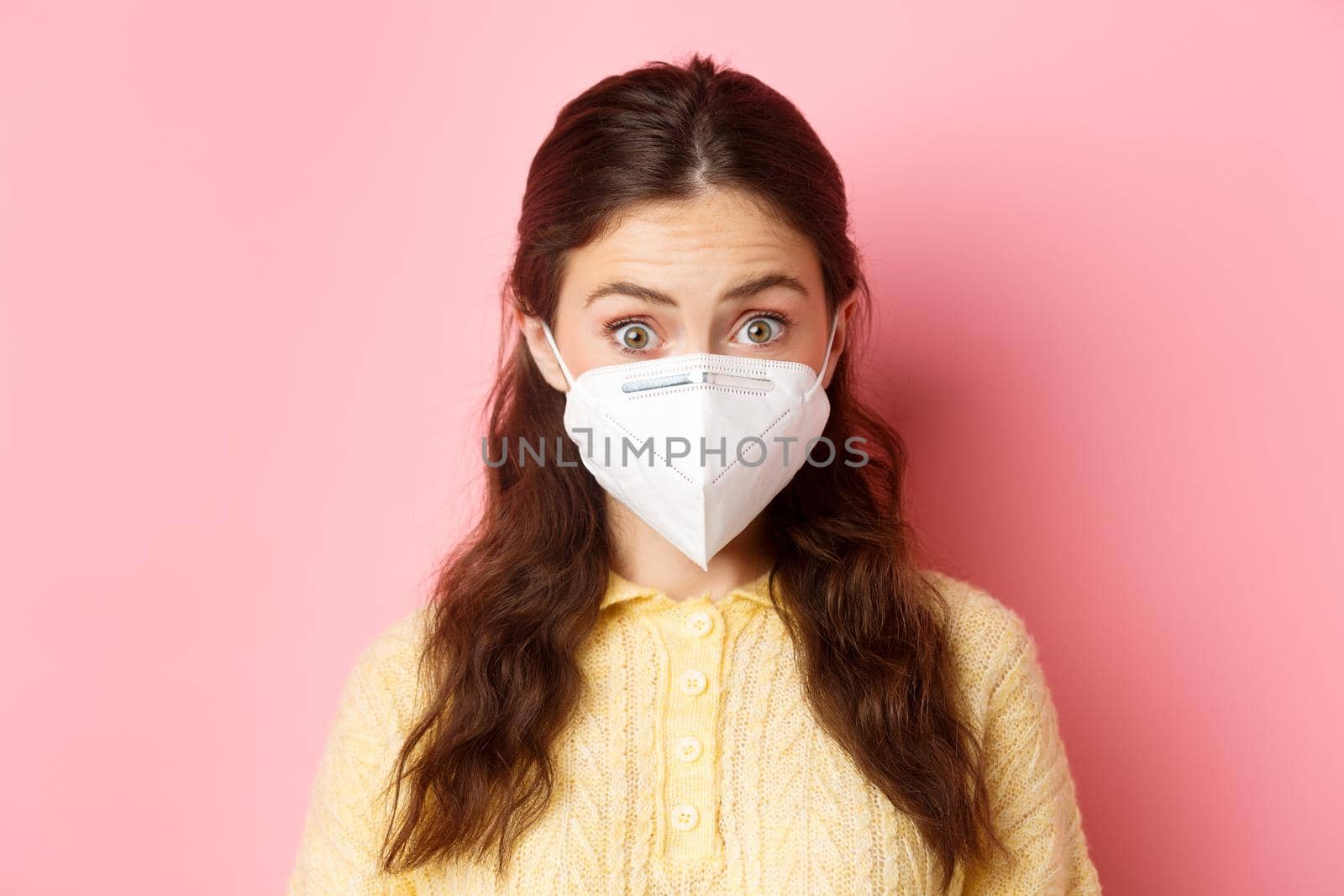 Preventive measures, health care concept. Close up portrait of woman in respirator raising eyebrows and looking surprised, hear interesting news, standing against pink background.