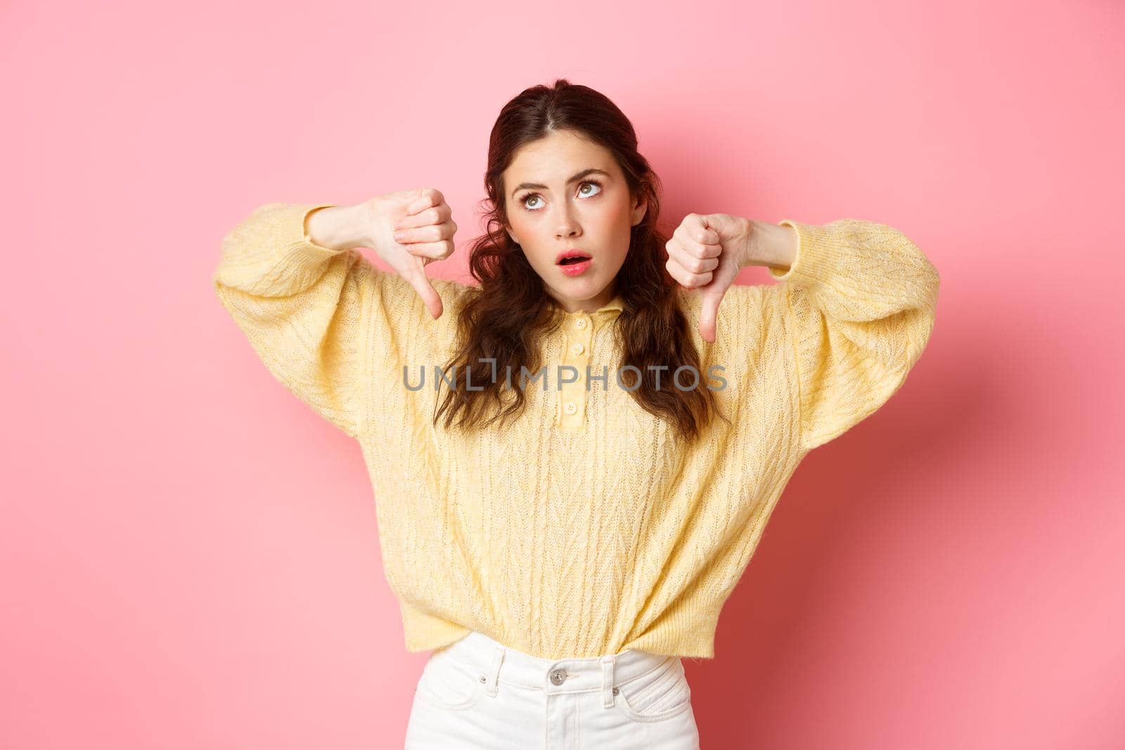 How lame. Displeased and indifferent young woman roll eyes, sighing and look aside, showing thumbs down with skeptical face, standing against pink background by Benzoix