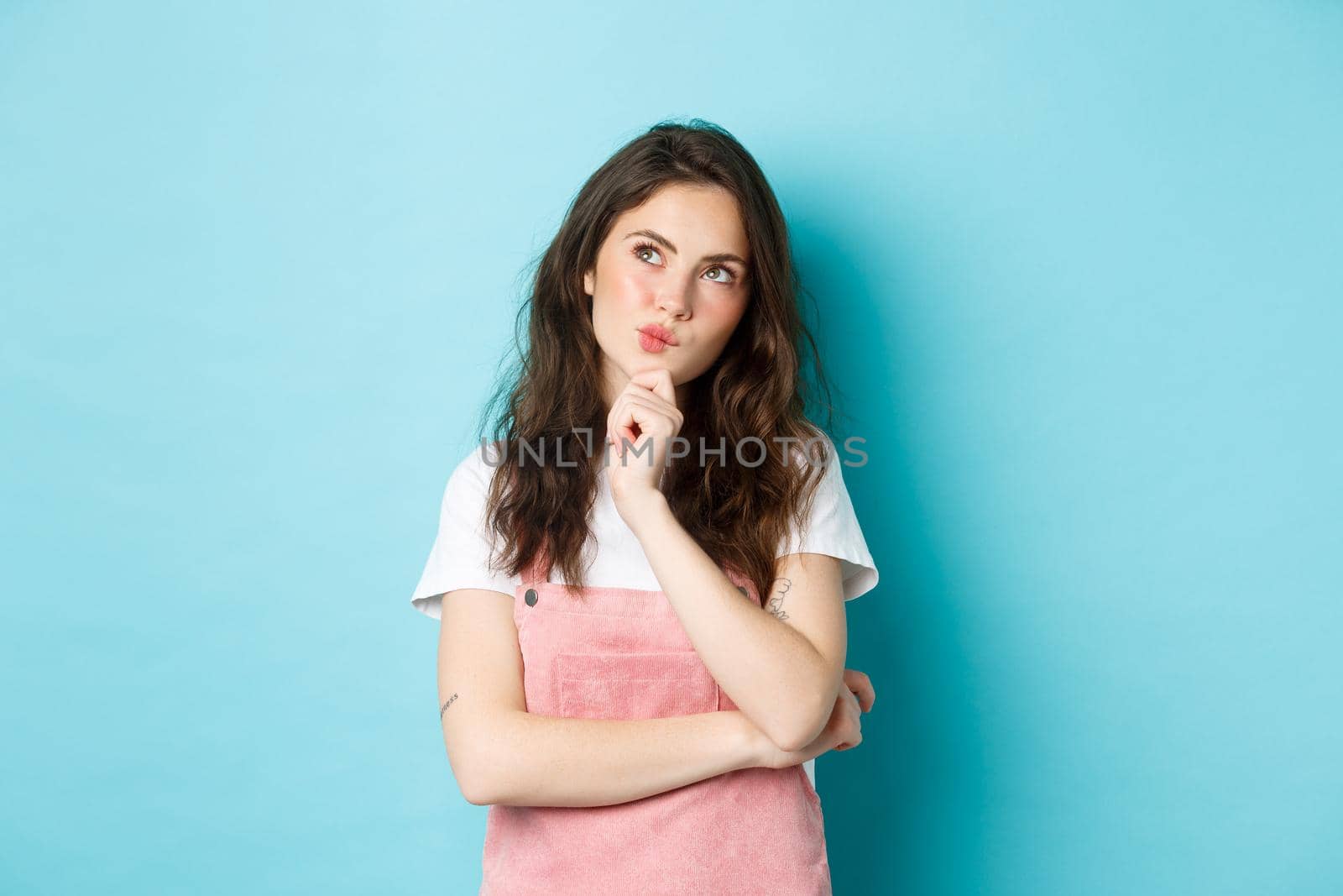 Thoughtful brunette woman making choice, holding hand on chin and thinking, looking at upper left corner with pensive face, consider something, standing over blue background by Benzoix