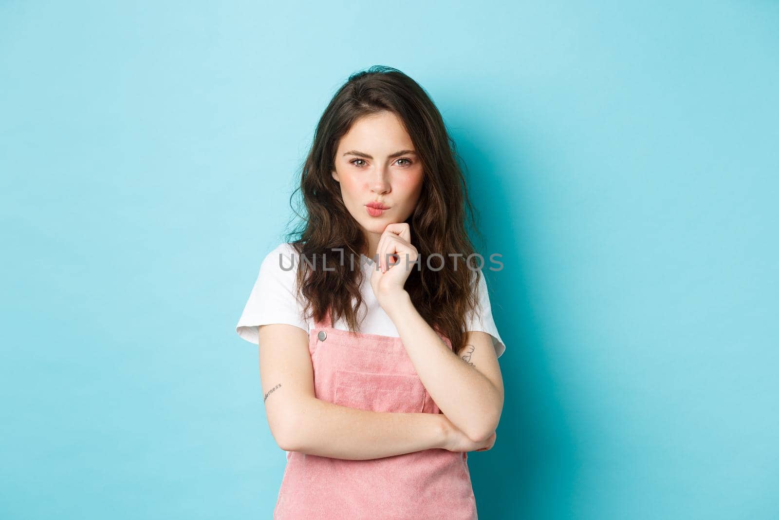 Thoughtful young teenage girl making assumption, look determined and pensive, planning something, staring at camera and thinking, standing over blue background.