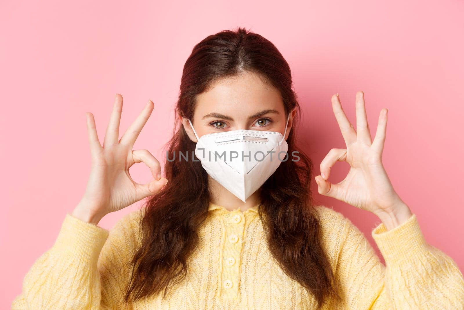 Preventive measures, health care concept. Close up of confident attractive woman, wearing medical respirator, looking determined and showing okay signs, standing against pink background by Benzoix
