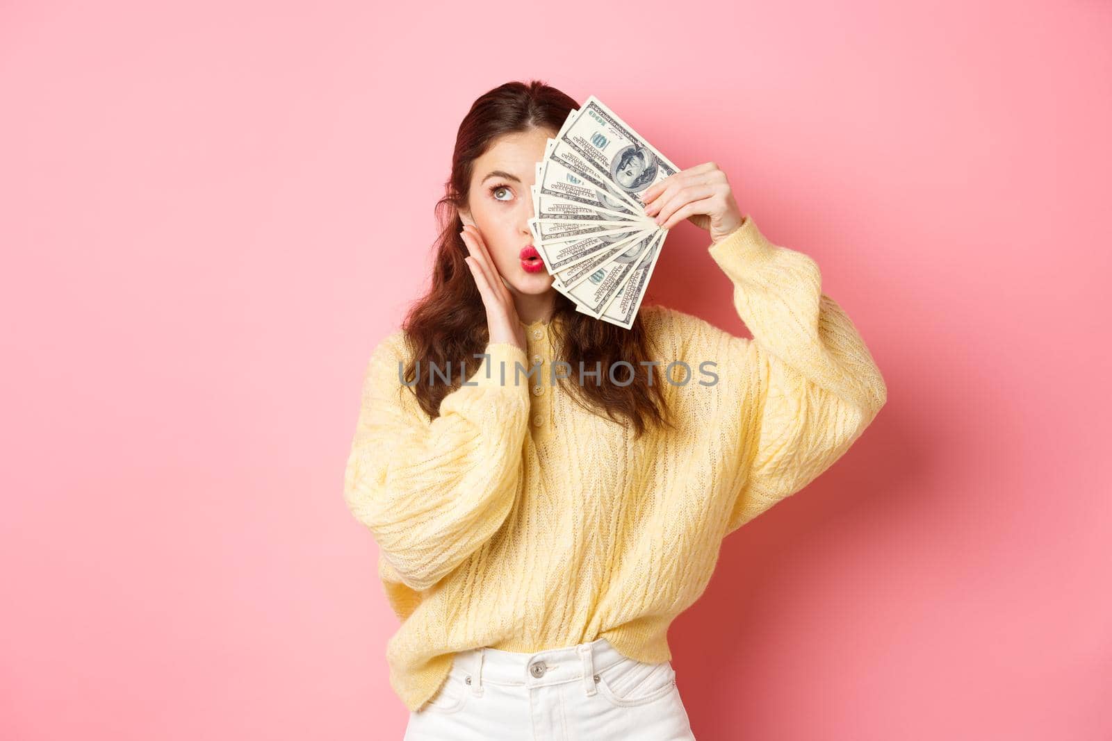 Attractive female model showing money, holding dollar bills on half of face, touching cheek and making kissing lips, standing against pink background.