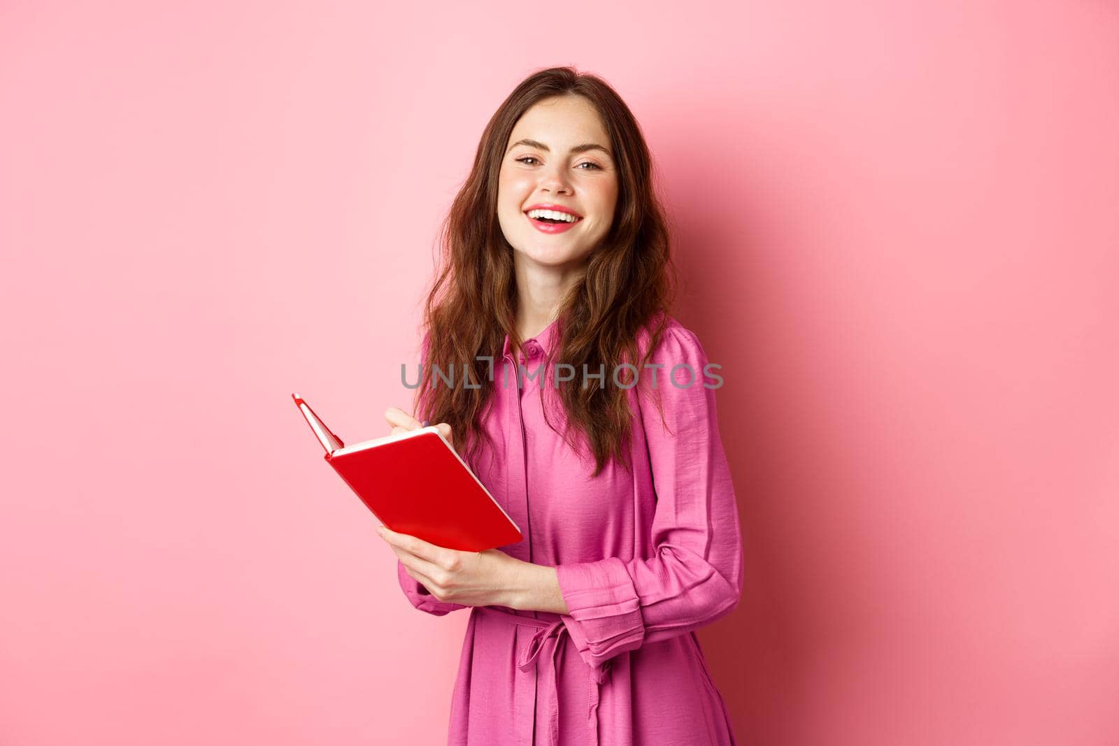 Beautiful young woman smiling, writing in notebook, holding planner or diary, standing against pink background.
