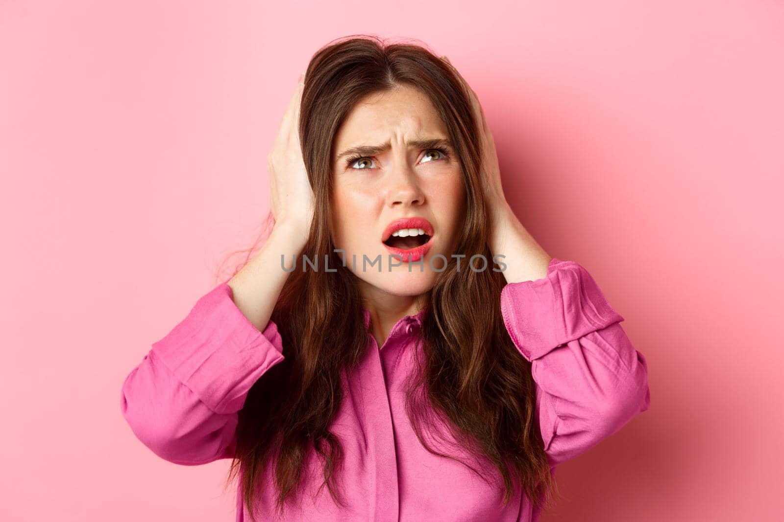 Close up of annoyed glamour girl covering ears with hands, looking bothered up at neighbours, cant stand loud music, standing over pink background.