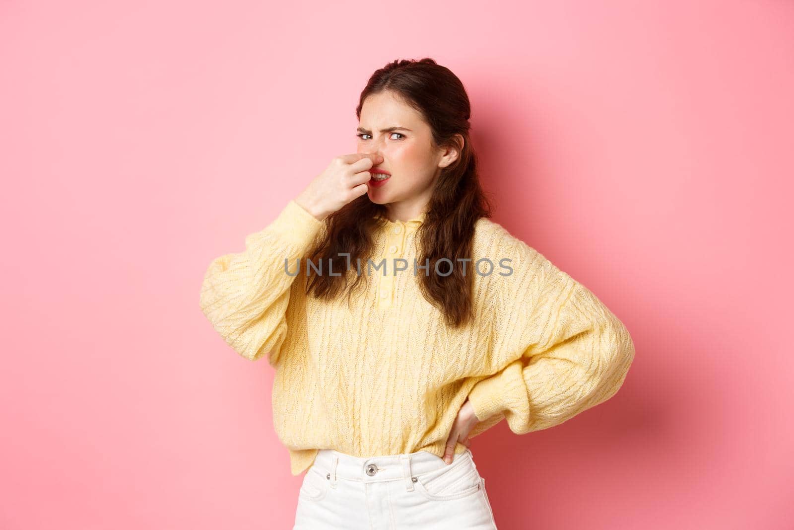 It stinks. Disgusted glamour girl shuts her nose from awful smell, look accusingly at camera, something reek bad, standing over pink background. Copy space