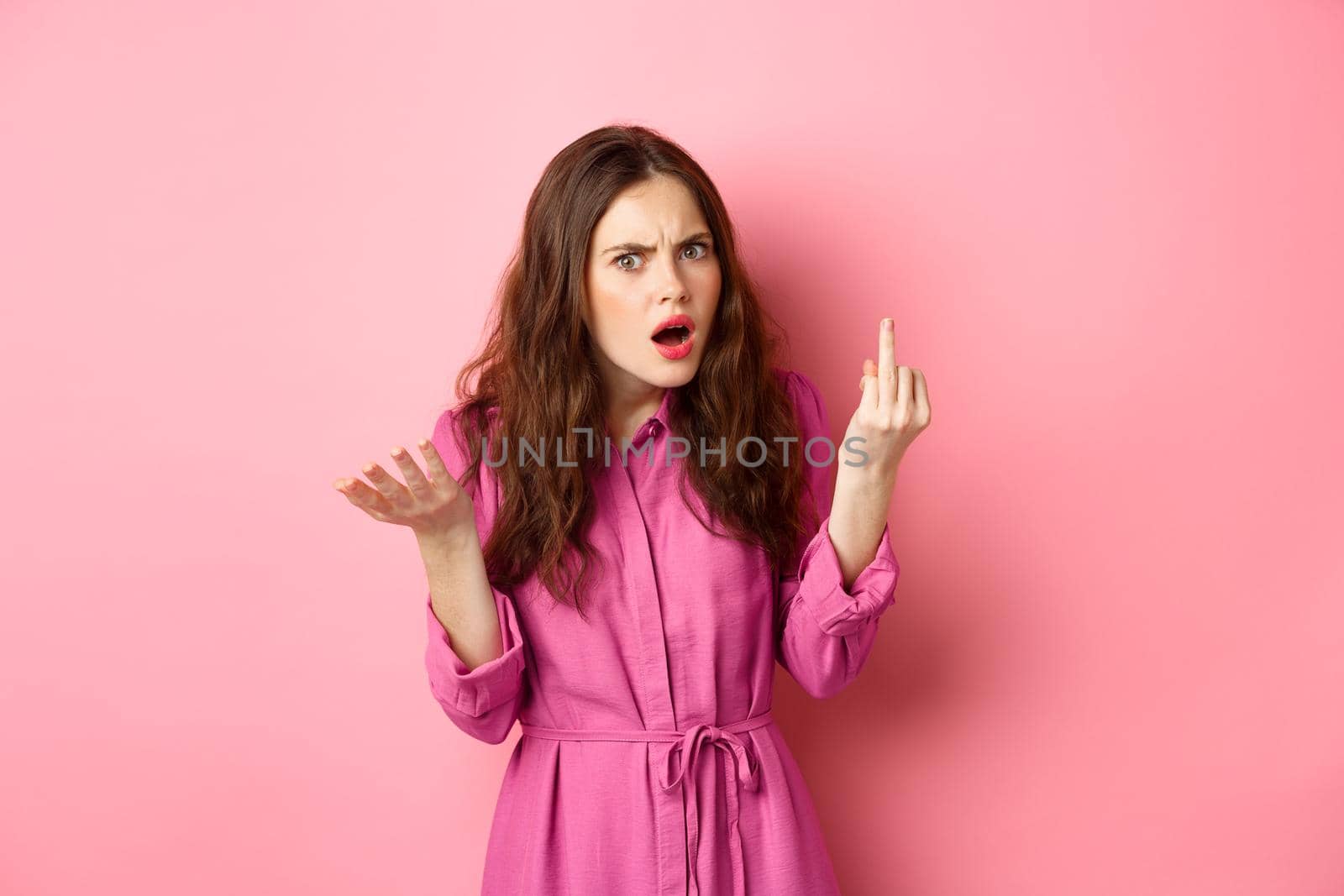 Annoyed girlfriend arguing about proposal, wants to get married, showing finger without engagement ring, standing confused against pink background.