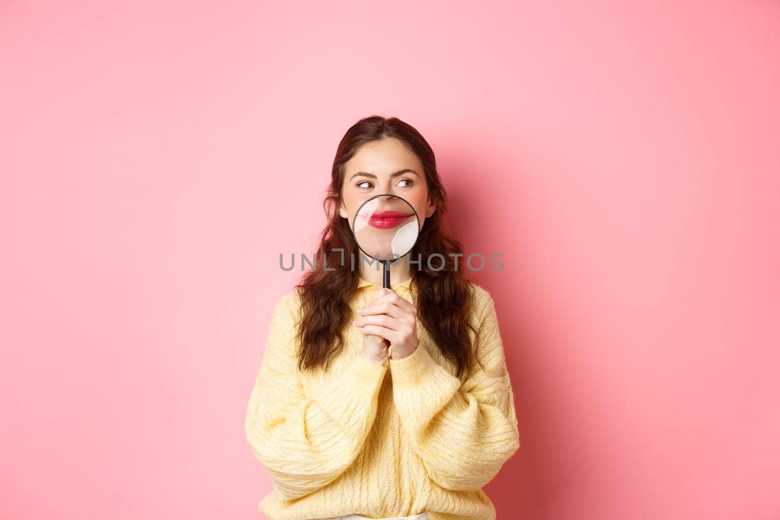 Image of coy attractive girl thinking of something interesting, showing her smile with magnifying glass near lips, standing against pink background, looking aside at promotional text.
