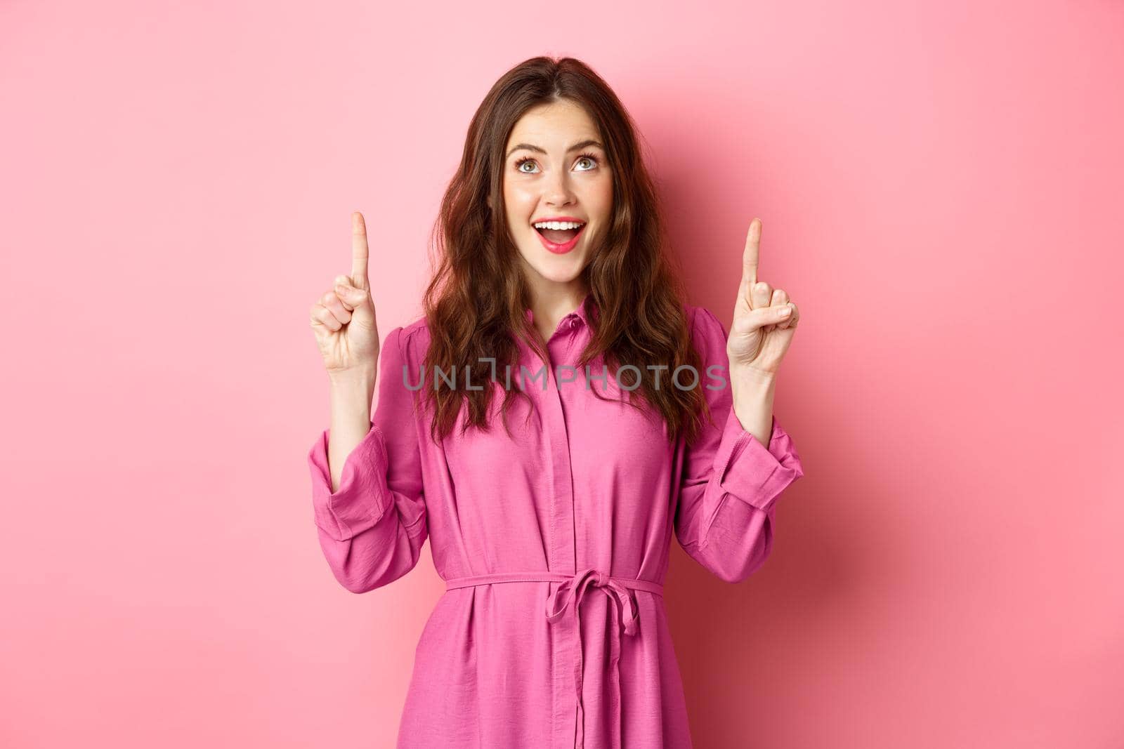 Beauty and fashion. Happy woman looking with amazement at copy space on top, pointing fingers up, smiling amazed, checking out promo offer, standing over pink background.