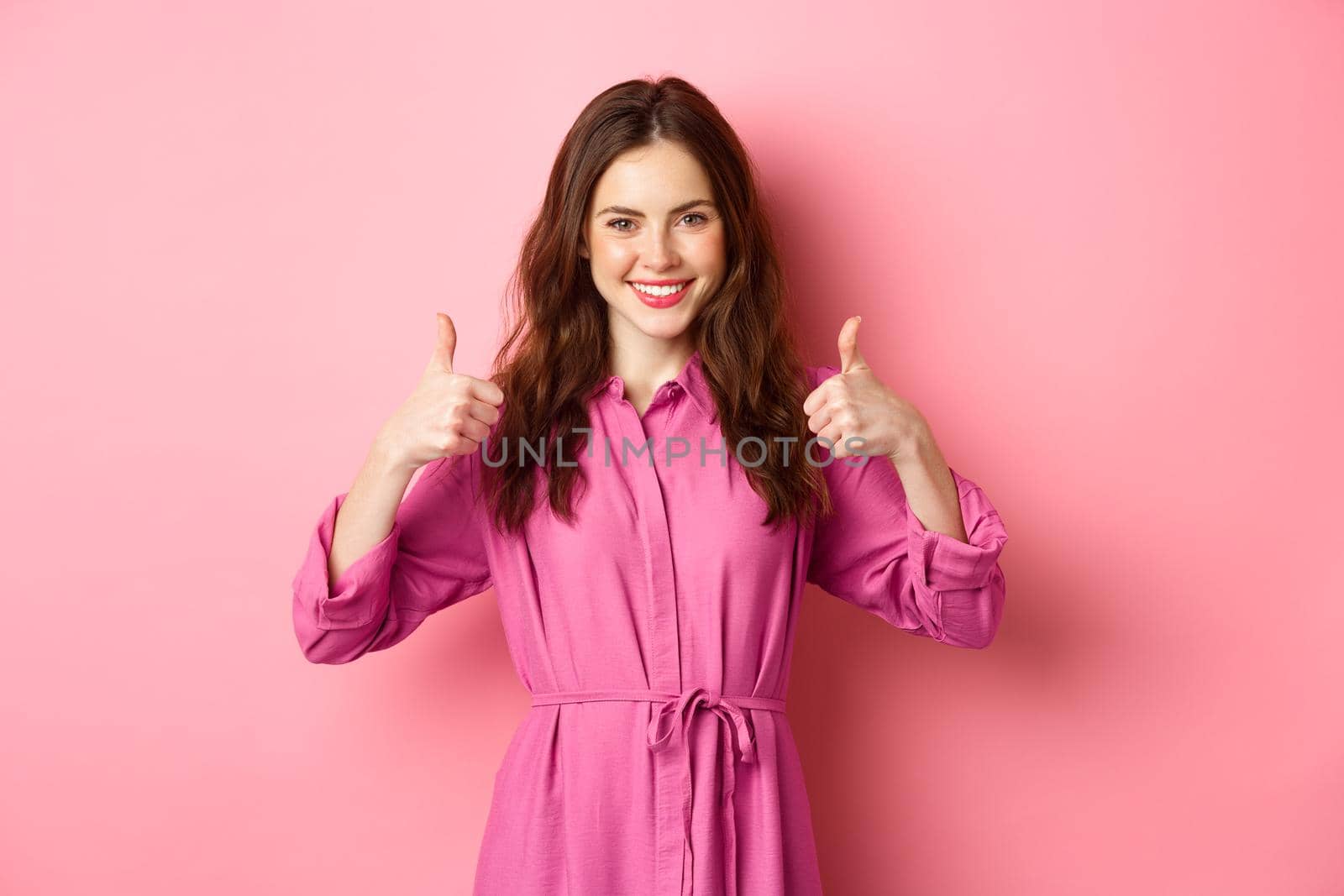 Satisfied female showing thumbs up in approval, say yes, agree and approve choice, praising you, standing against pink background.
