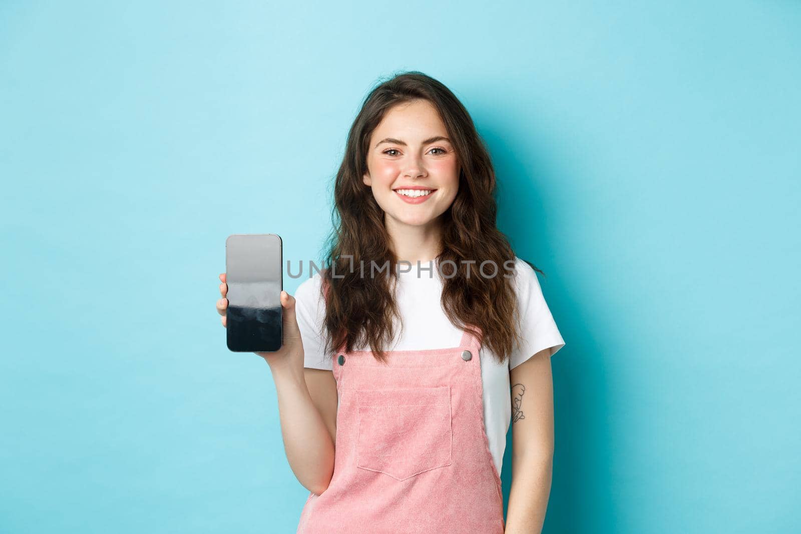 Portrait of smiling beautiful woman showing her smartphone screen and looking pleased, recommending application or online page, standing over blue background.