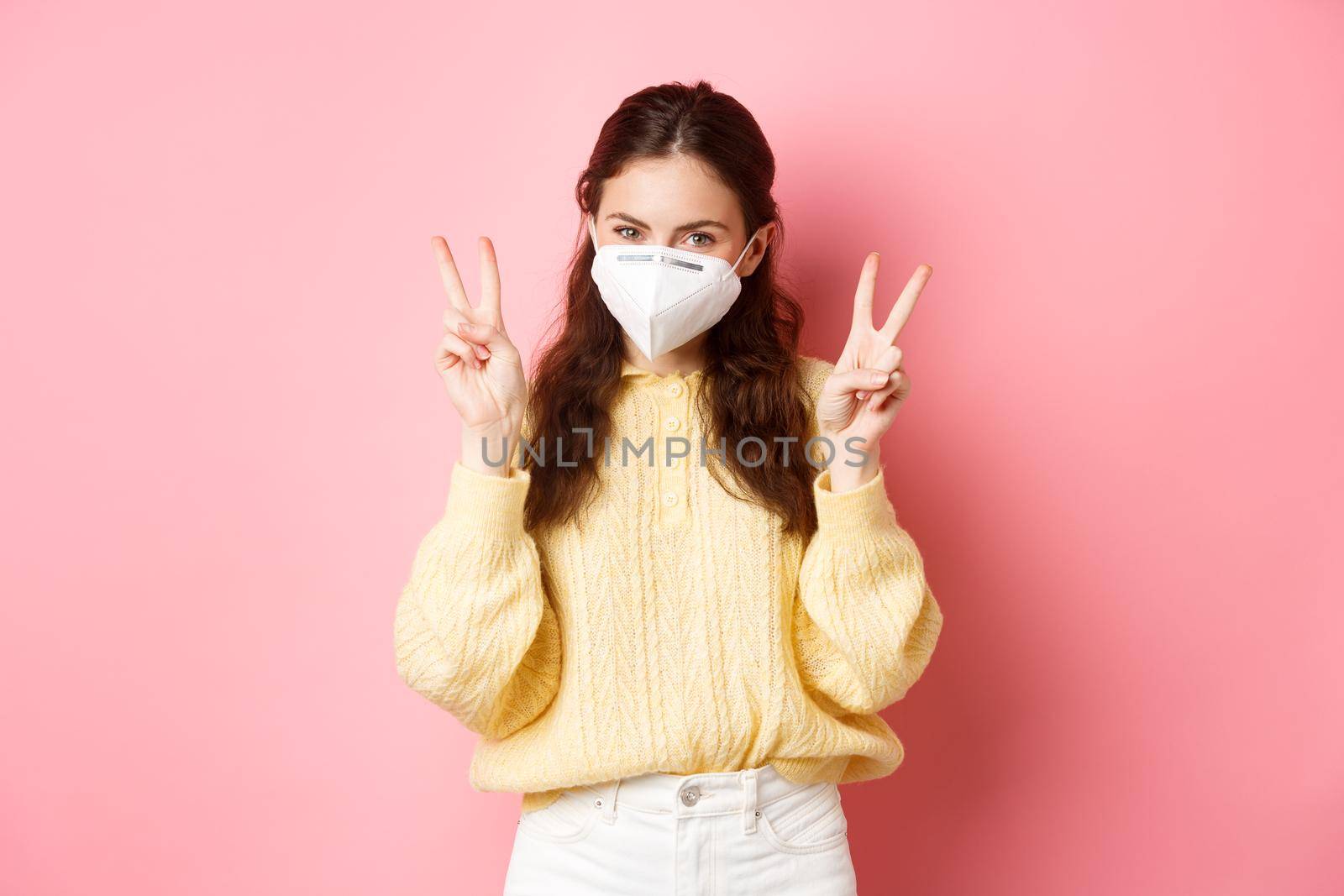 Covid-19, lockdown and pandemic concept. Cute glamour girl wears medical respirator for going outdoors during quarantine, shows peace v-sign, stands against pink background by Benzoix
