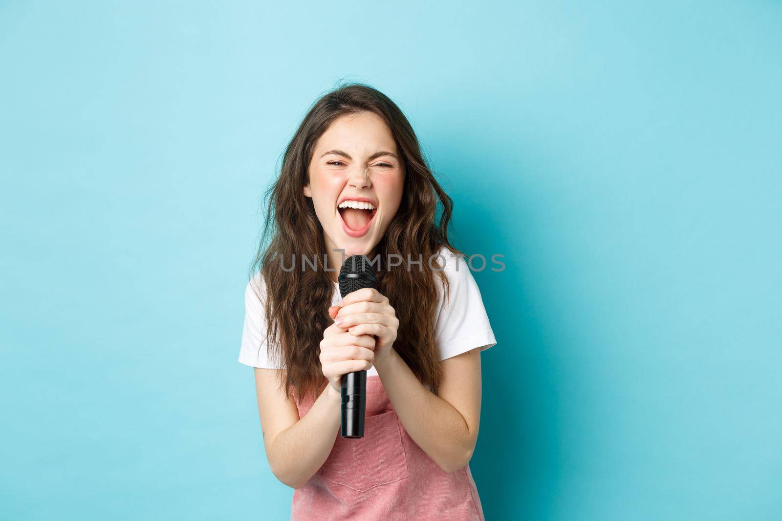 Excited pretty girl singing karaoke, holding microphone and smiling happy, standing over blue background.