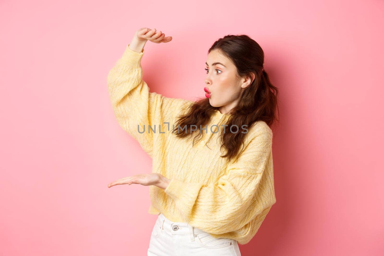 Portrait of brunette girl holds big empty space and stare amazed at place for your product or logo, showing large box gesture, standing against pink background.