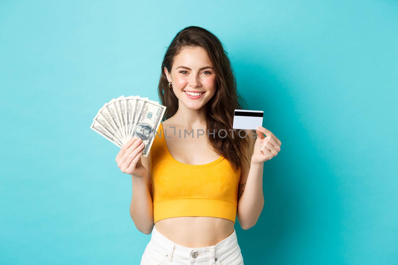 Portrait of young woman with smiling face, showing plastic credit card and money, standing against blue background.