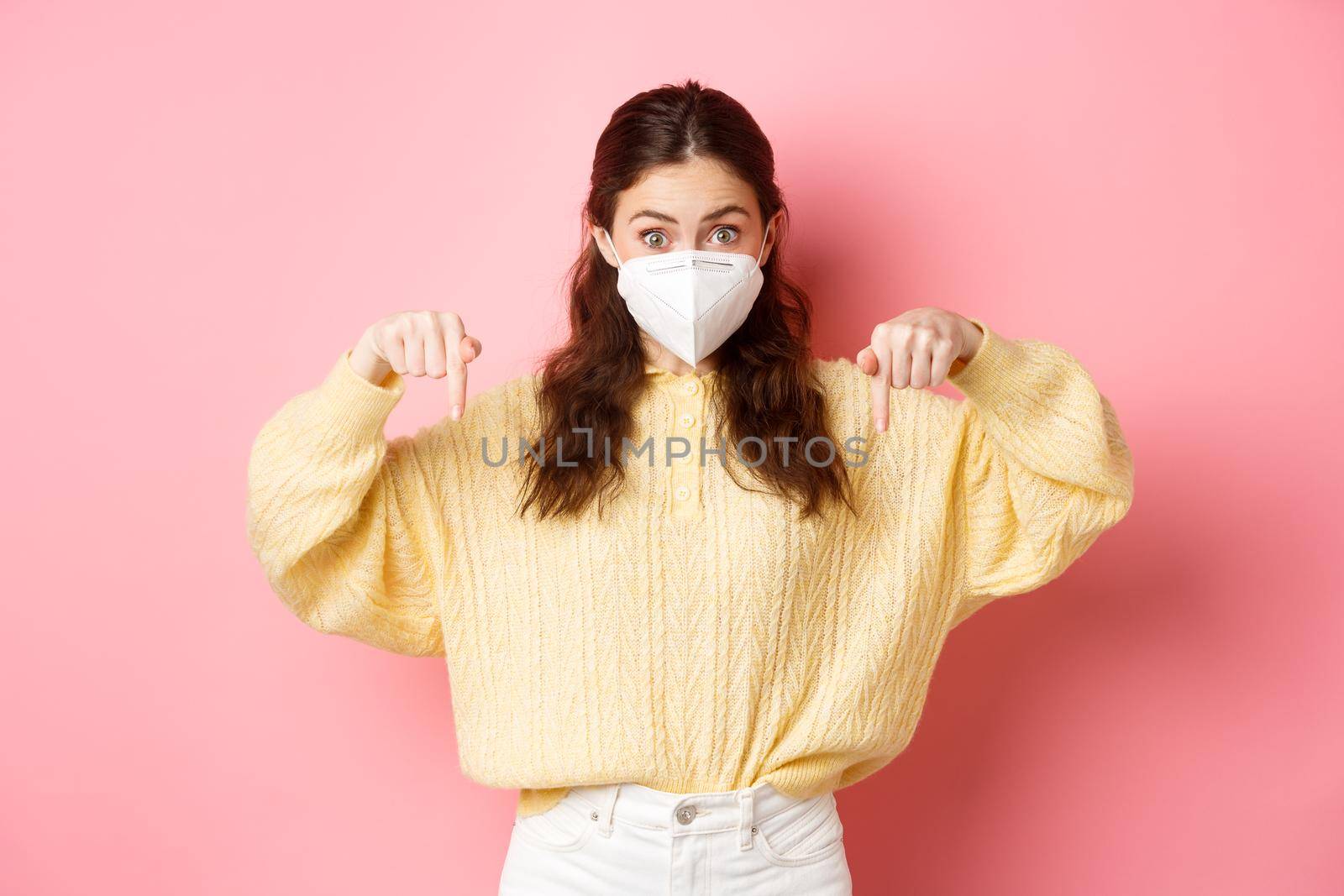 Preventive measures, health care concept. Excited woman in medical respirator pointing fingers down at copyspace, showing promo, standing against pink background.