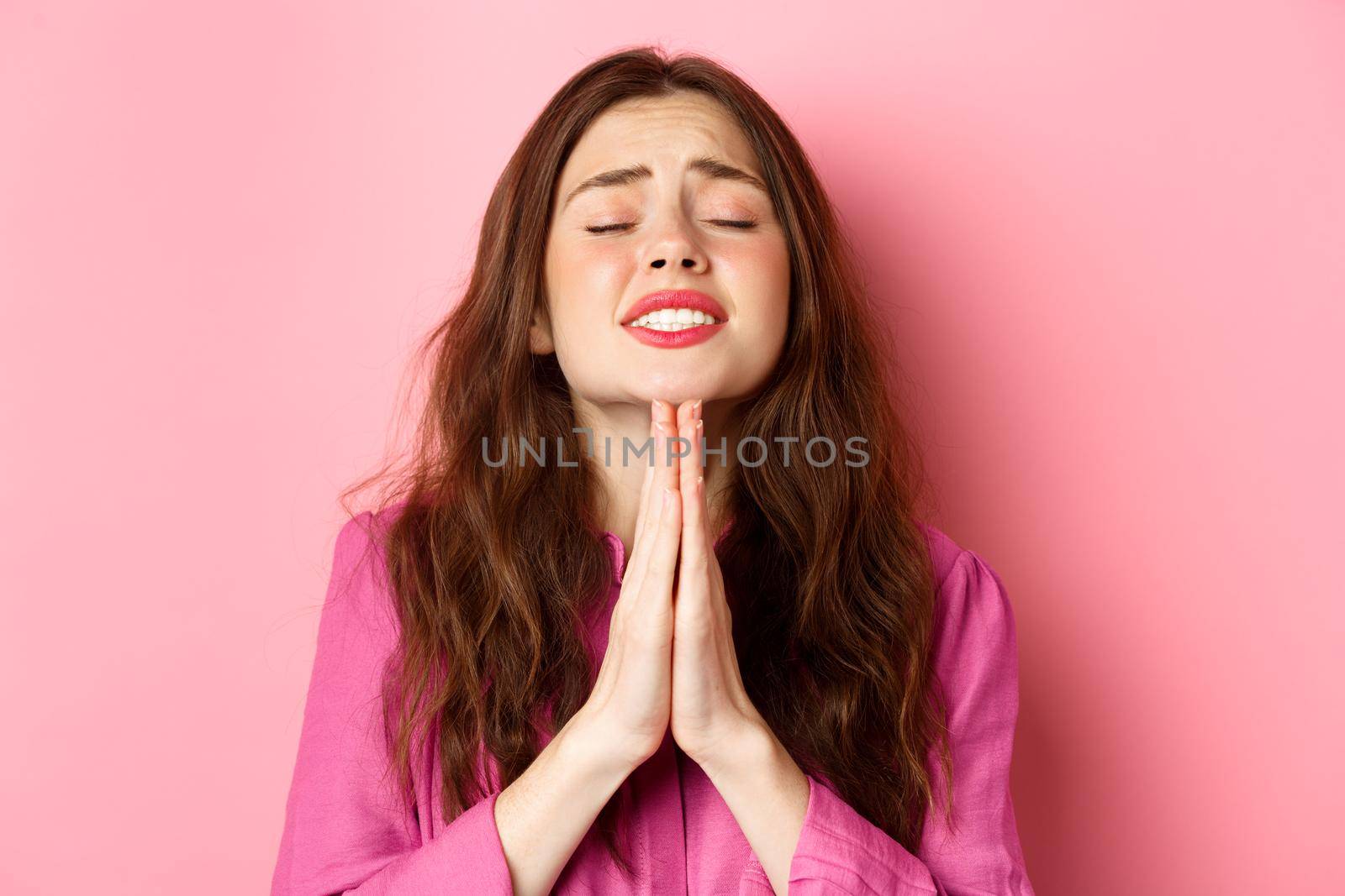 Image of hopeful young woman in need, praying, begging for help, standing over pink background in supplication pose.