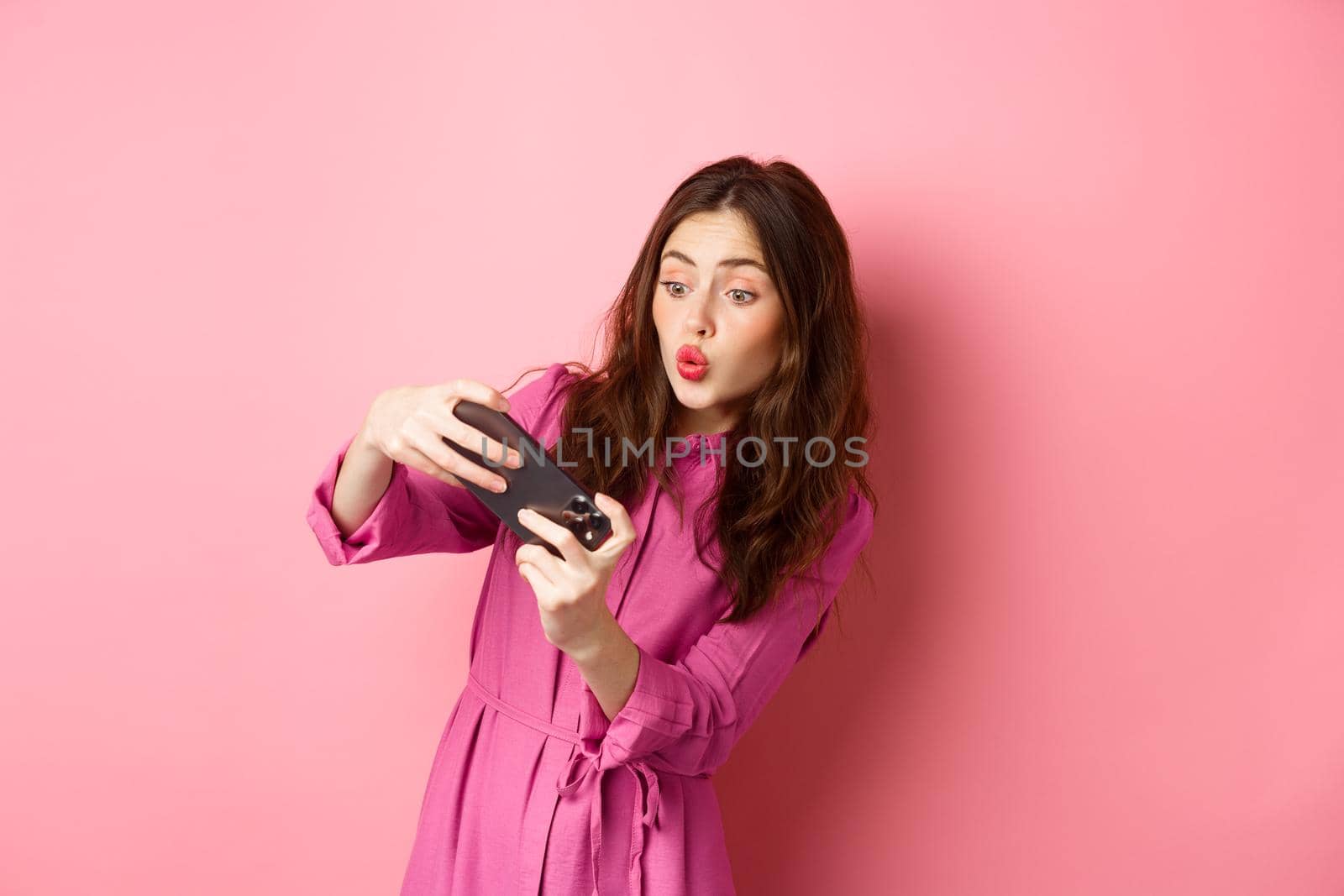 Technology concept. Carefree young woman tilt body, holding smartphone horizontally, playing video game on phone, standing against pink background by Benzoix