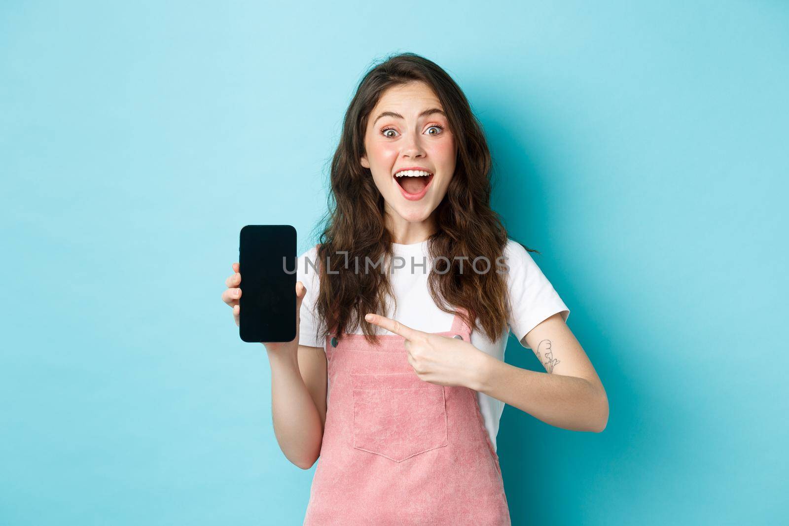 Wow check this out. Excited pretty girl pointing finger at phone screen, showing logo or store advertisement on smartphone, standing against blue background by Benzoix