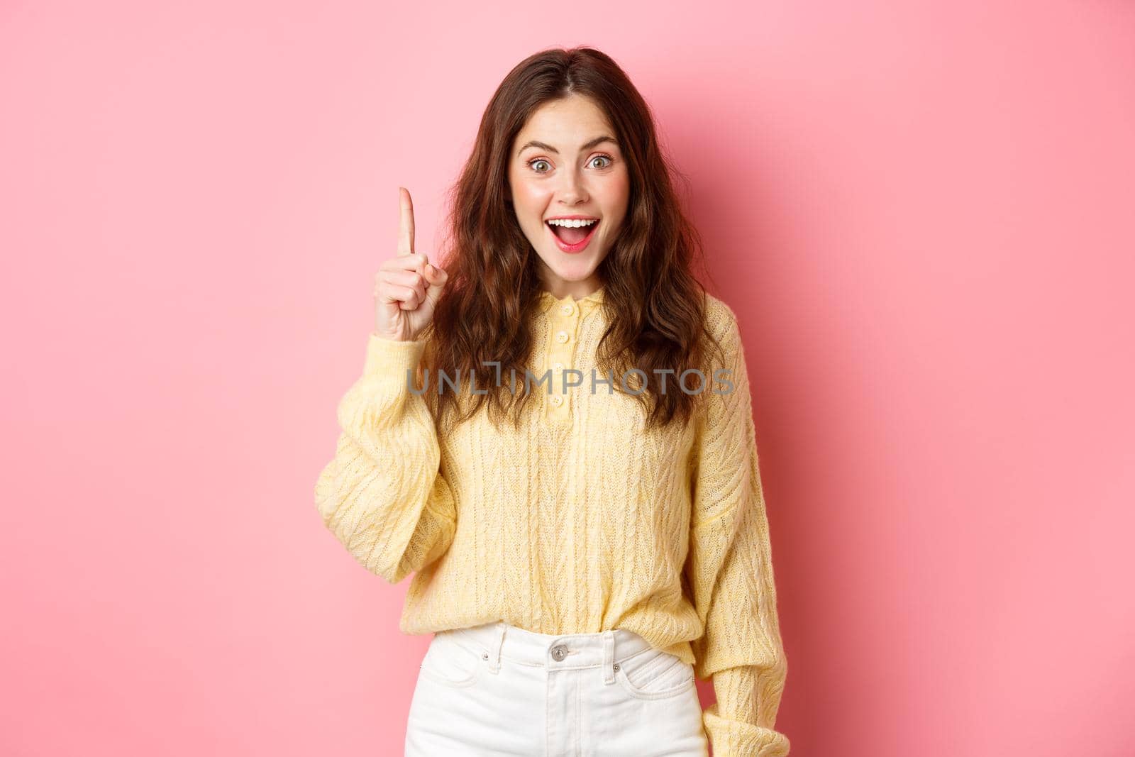 Young excited woman in stylish outfit, raising finger up, pitching an idea, saying suggestion, ahve interesting plan, standing over pink background.
