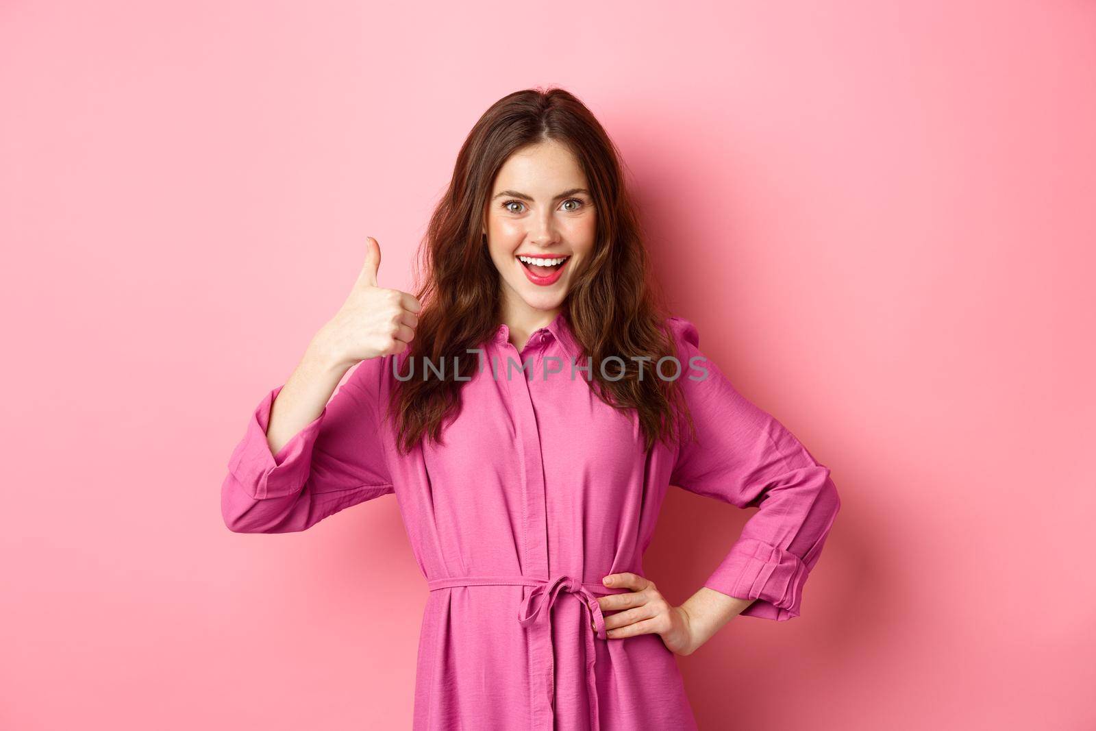 Young attractive woman saying yes, showing thumbs up sign in approval, smiling confident and satisfied, standing over pink background.