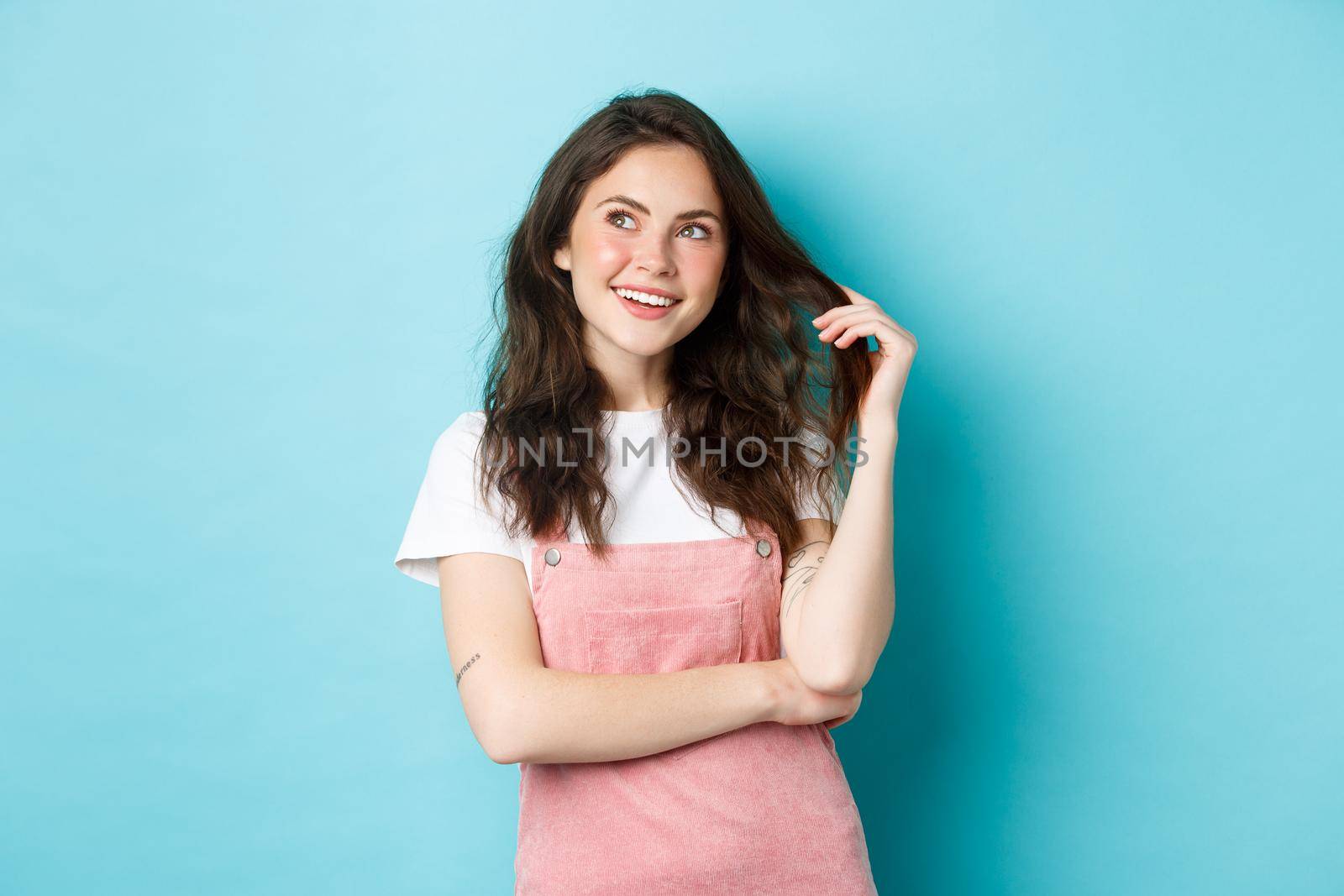 Young girl looking dreamy and playing with glowing curly hair, smiling pensive, imaging summer holidays or shopping, standing against blue background.