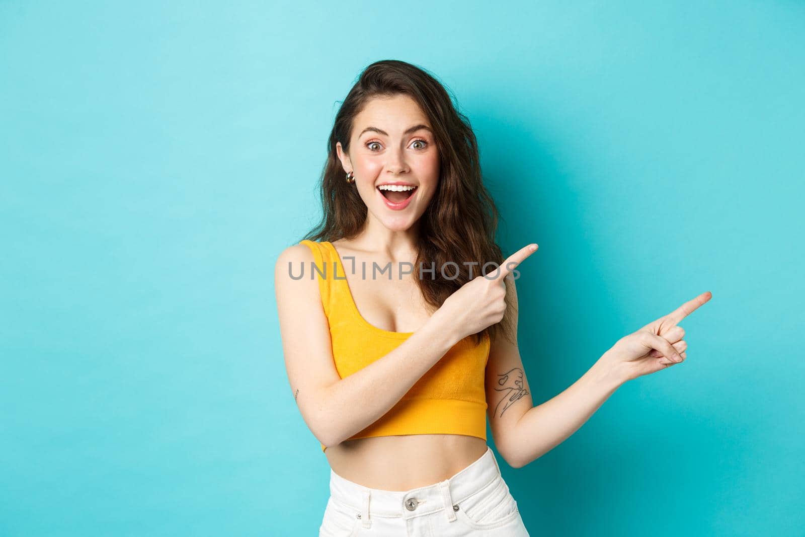 Summer lifestyle concept. Cheerful stylish woman showing direction, pointing fingers right, that way gesture, standing over blue background.