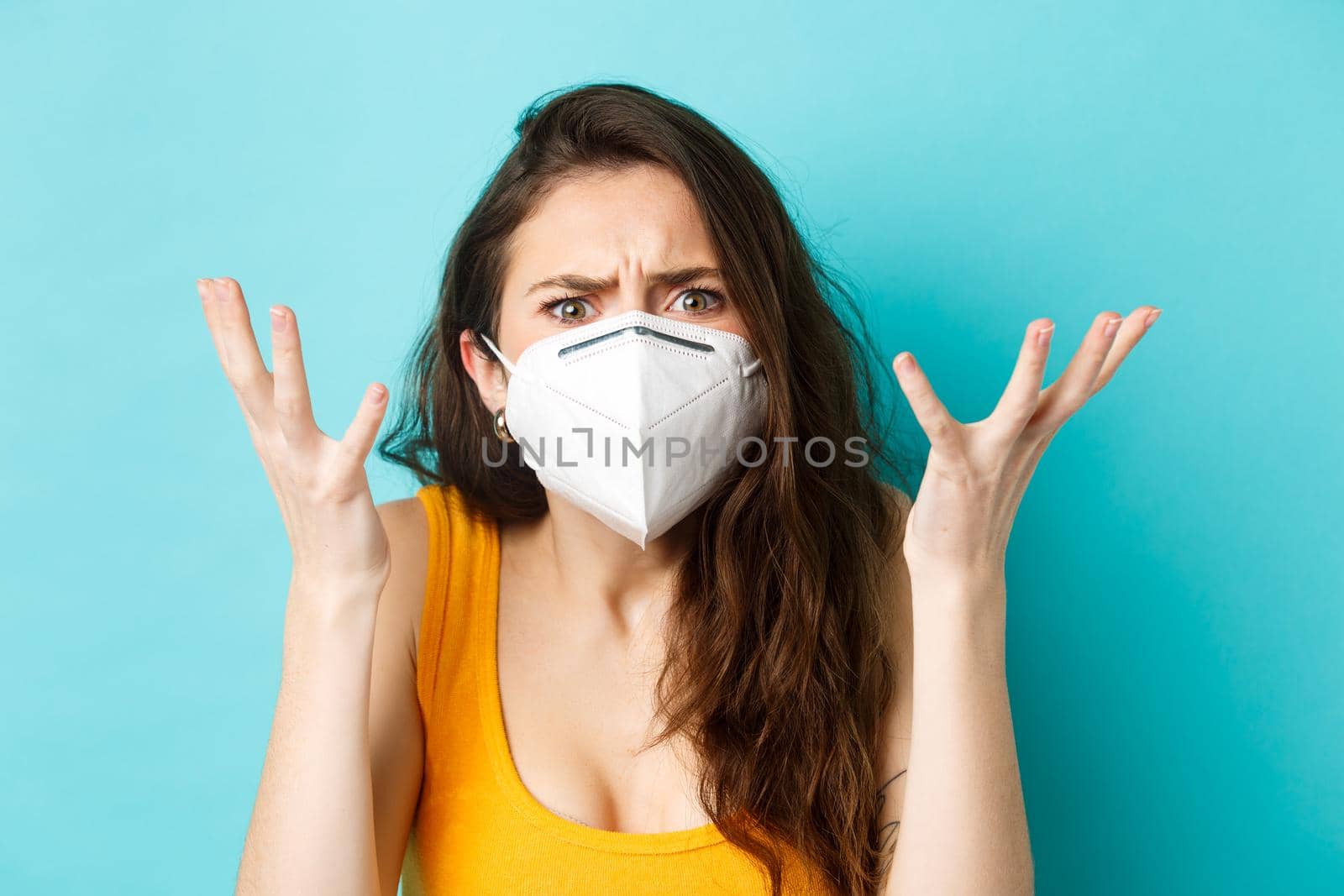 Corona, pandemic and healthcare concept. Annoyed and confused beautiful woman arguing, wearing respirator, shaking hands and looking displeased at camera, blue background.
