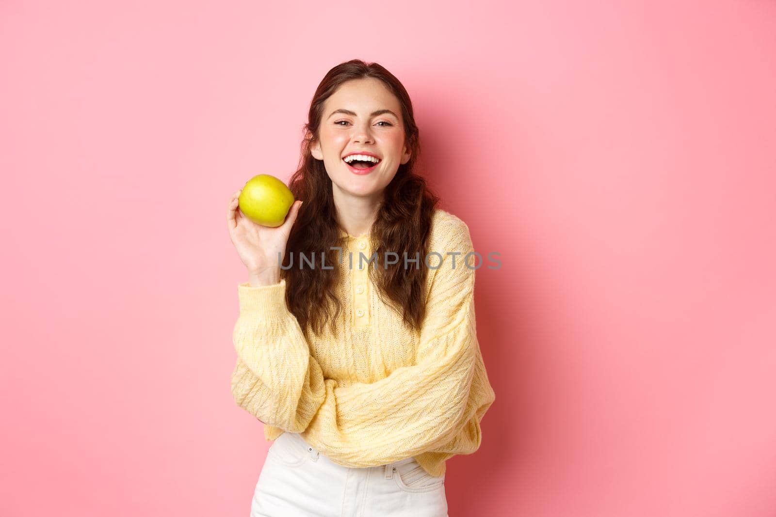 Healthy diet, people and lifestyle concept. An apple day keeps doctor away, girl holding delicious fruit and smiling happy at camera, standing against pink background.