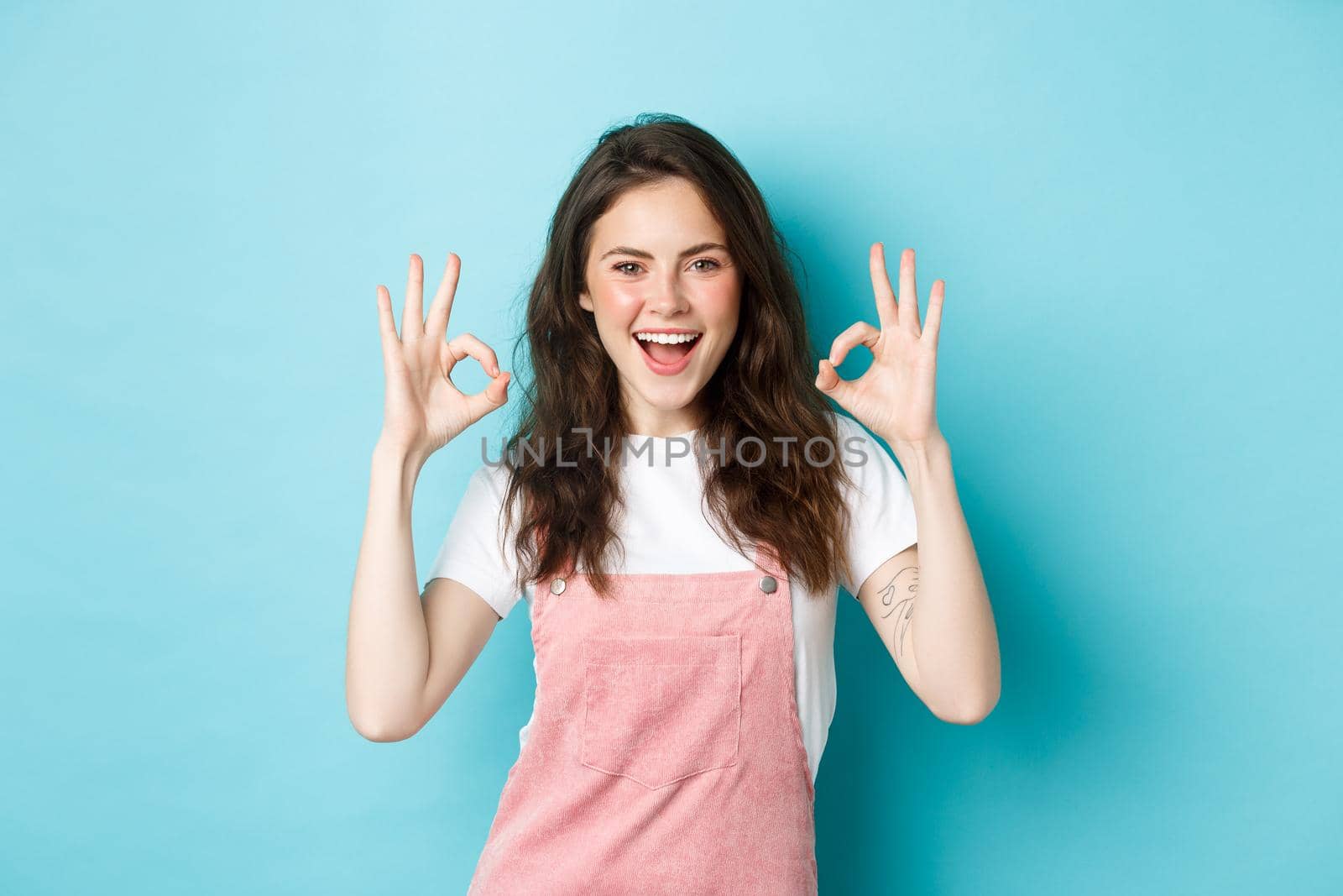 Very well, alright. Excited smiling brunette guy say yes, showing okay signs and agree with you, praising good job, standing happy against blue background.