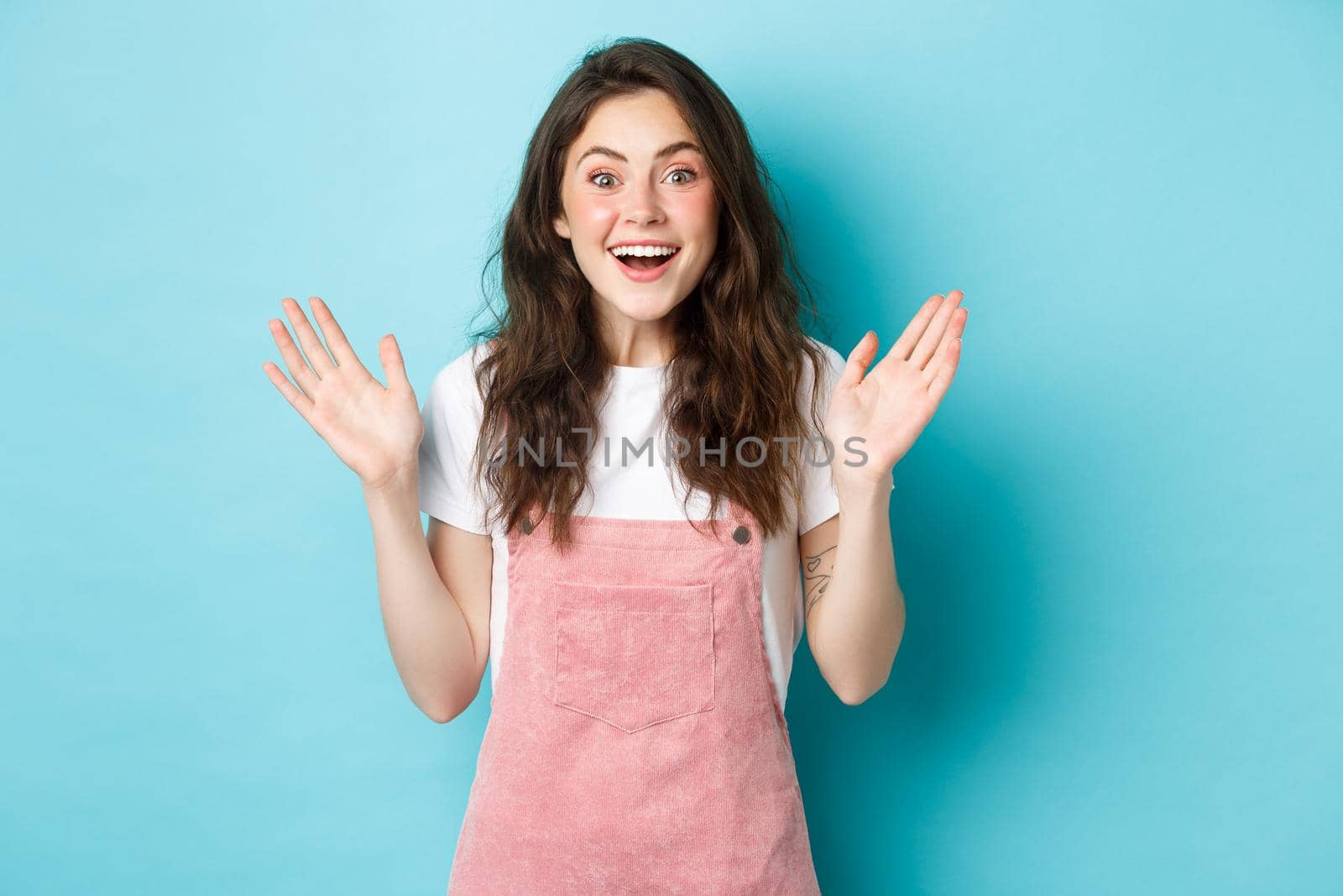 Portrait of beautiful teenage girl laughing and smiling, look surprised and clap hands from amazement and joy, staring amazed at camera, standing against blue background.