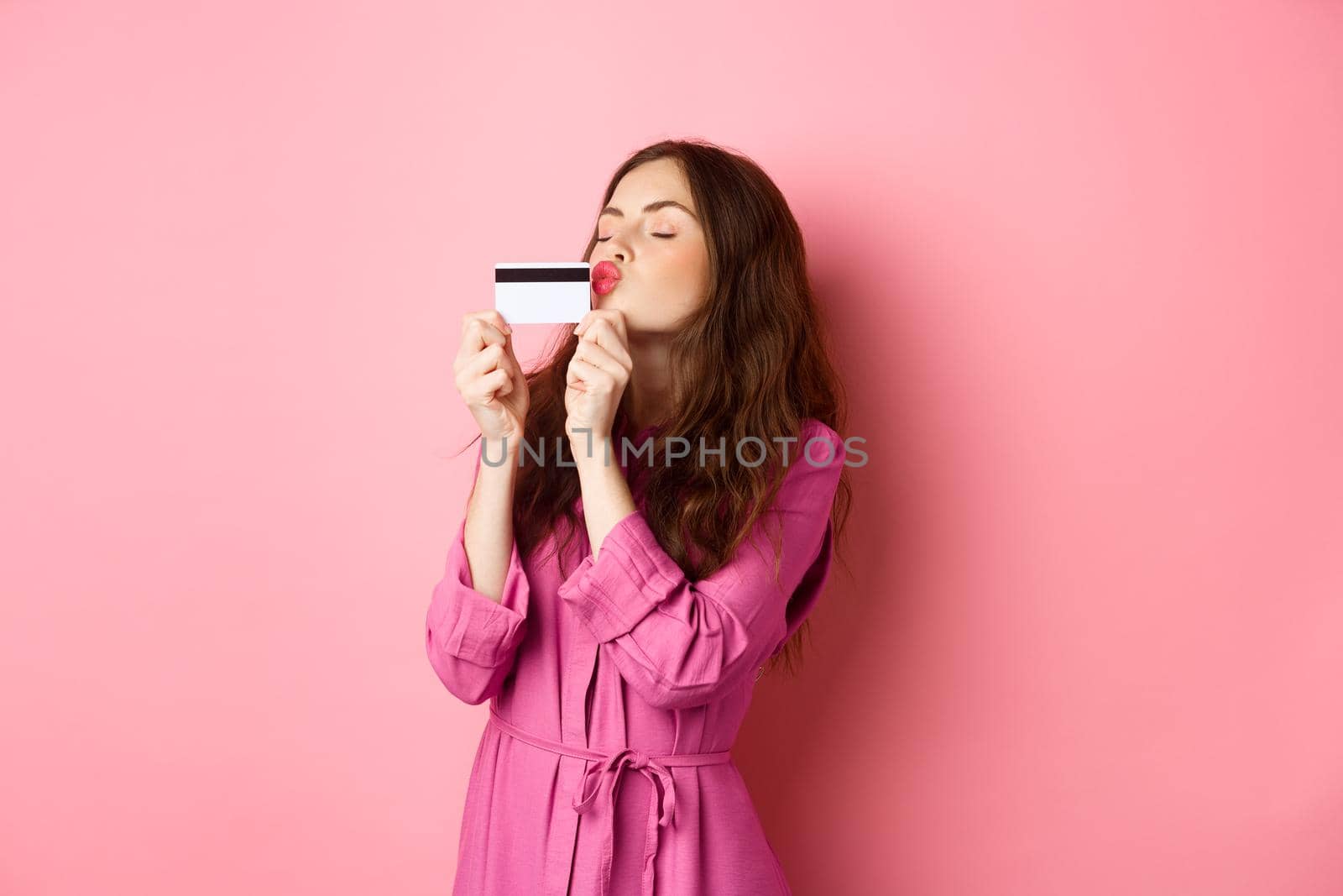 Image of young beautiful woman kissing her credit card, going on shopping, wasting money in stores, standing against pink background. Copy space