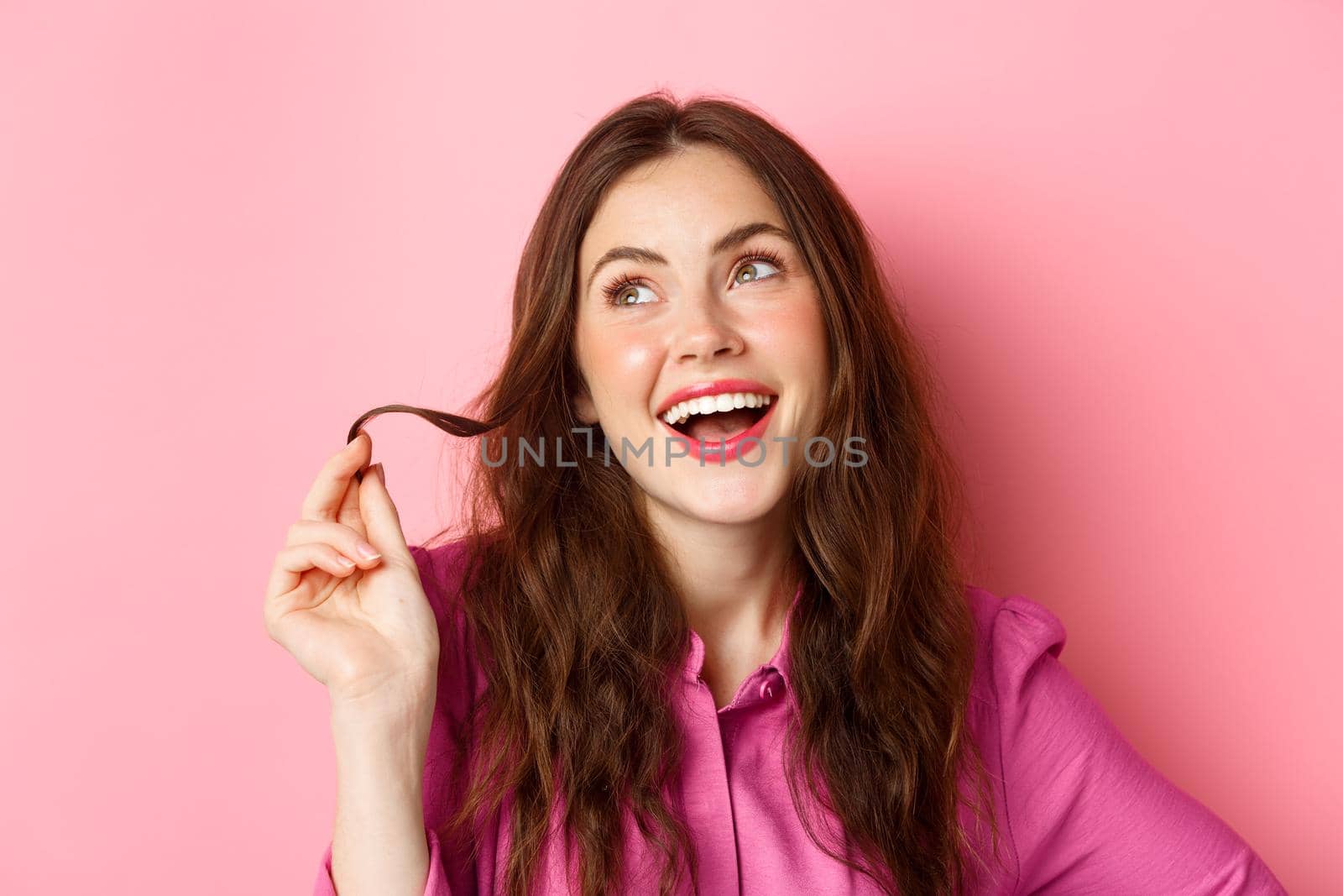 Fashion and beauty concept. Stylish young woman with make up and hairstyle, playing with hair strand and looking aside with coquettish, thoughtful smile, standing against pink background.