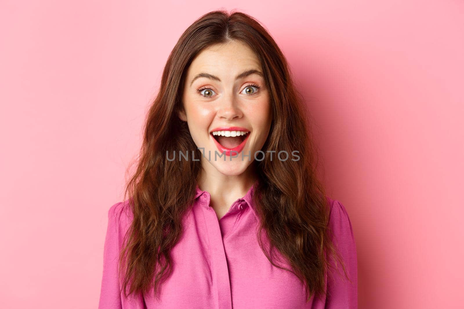 Close up portrait of attractive woman gasping, looking surprised and amazed at camera, hear amazing news, pink background.
