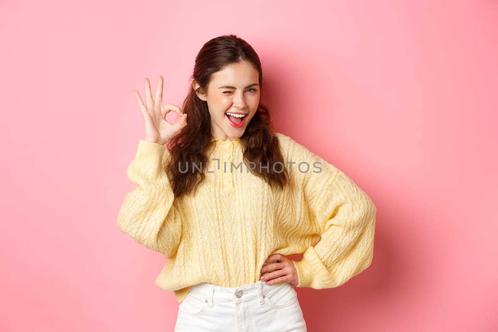Cheeky young woman winking, showing okay sign, give her approval, like and approve good thing. Girl make OK gesture to give permission, say yes, standing over pink background.