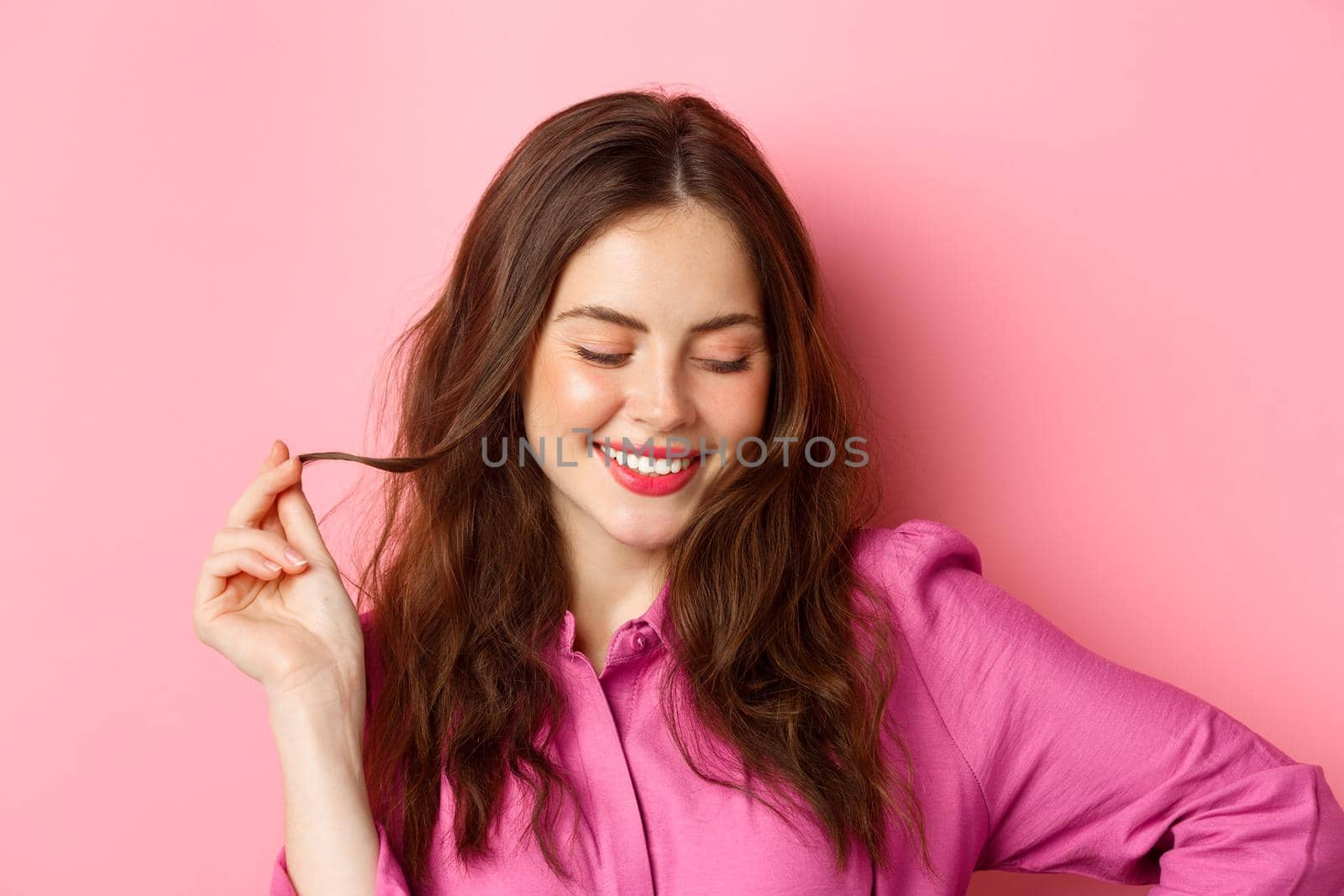 Fashion and beauty concept. Close up of flirty, happy young woman blushing, making romantic cute smile and looking down, playing with hair as flirting, standing against pink background.