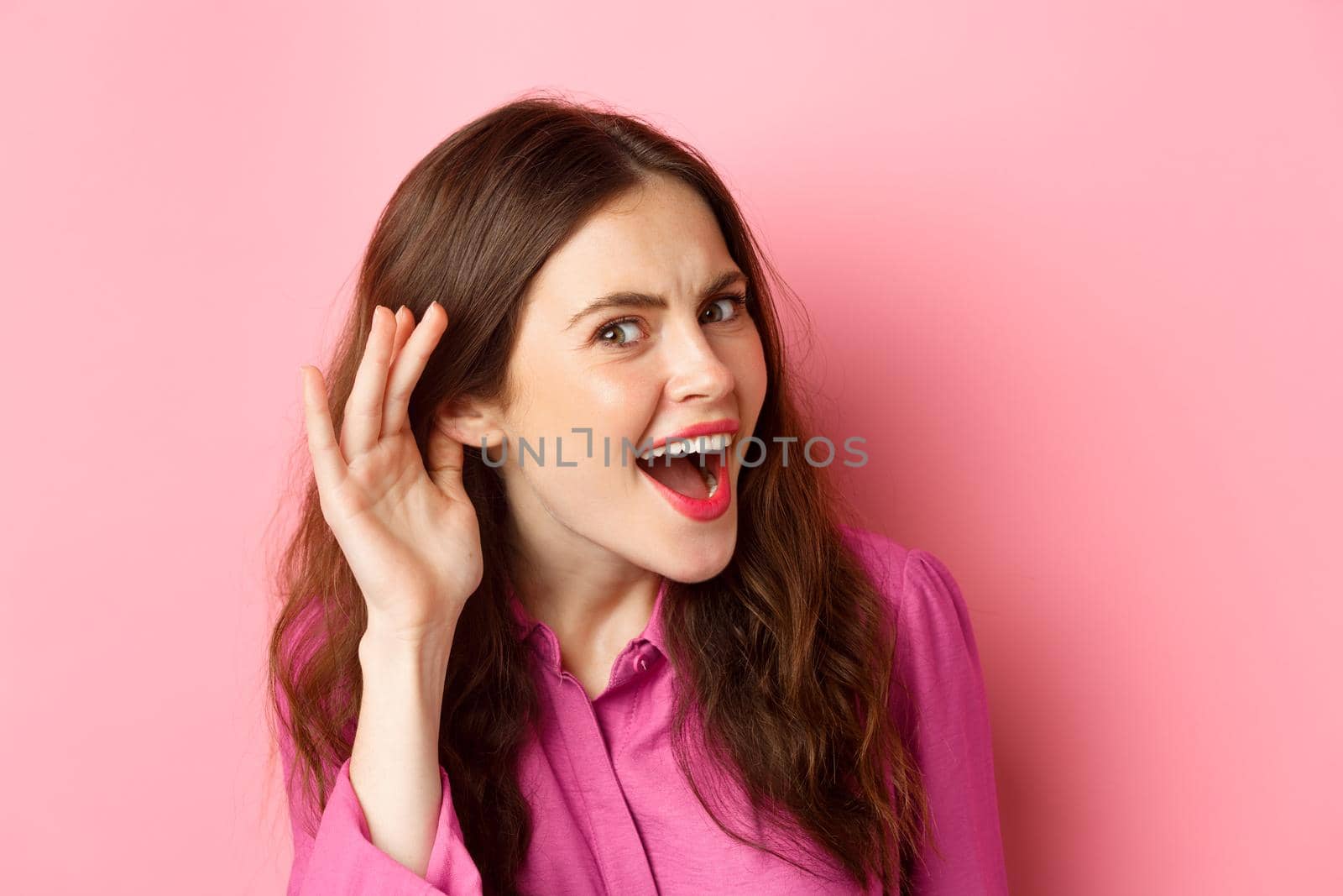 Close up of excited young woman overhear juice rumor, lean closer to camera with hand near ear, eavesdropping, standing over pink background.